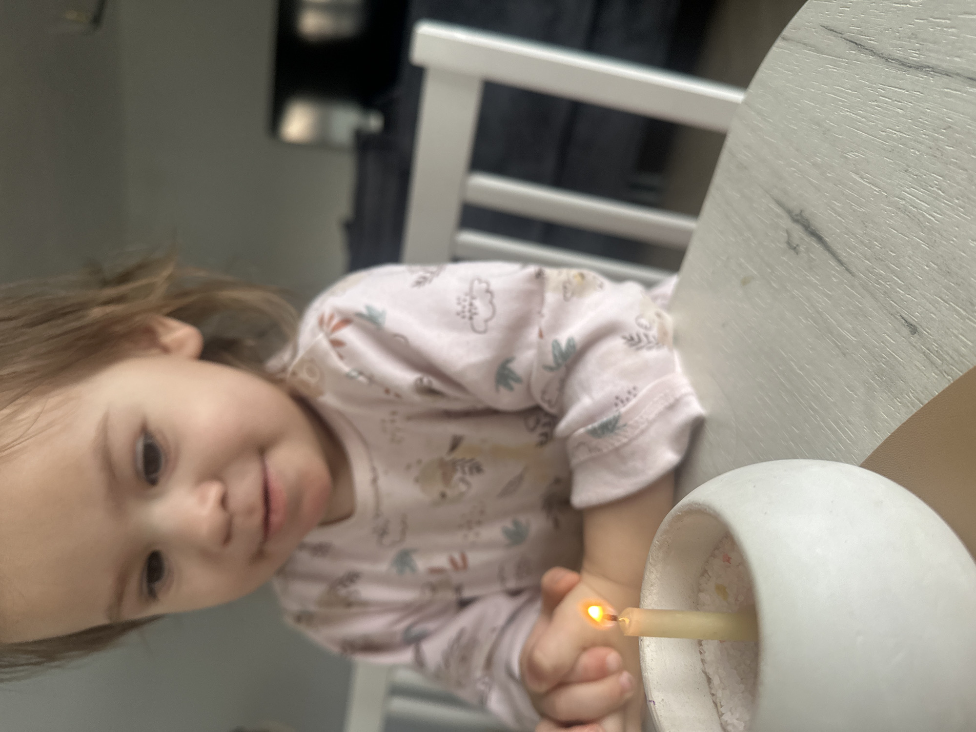 A young child wearing a pink patterned top sits at a table. The child is looking towards the camera. A lit candle sits in a white ceramic candle holder filled with white granular material. The table has a light-colored, faux-wood grain surface. The back of a white chair is visible behind the child. The location Kharnavy, Russia is not discernible from the image.
