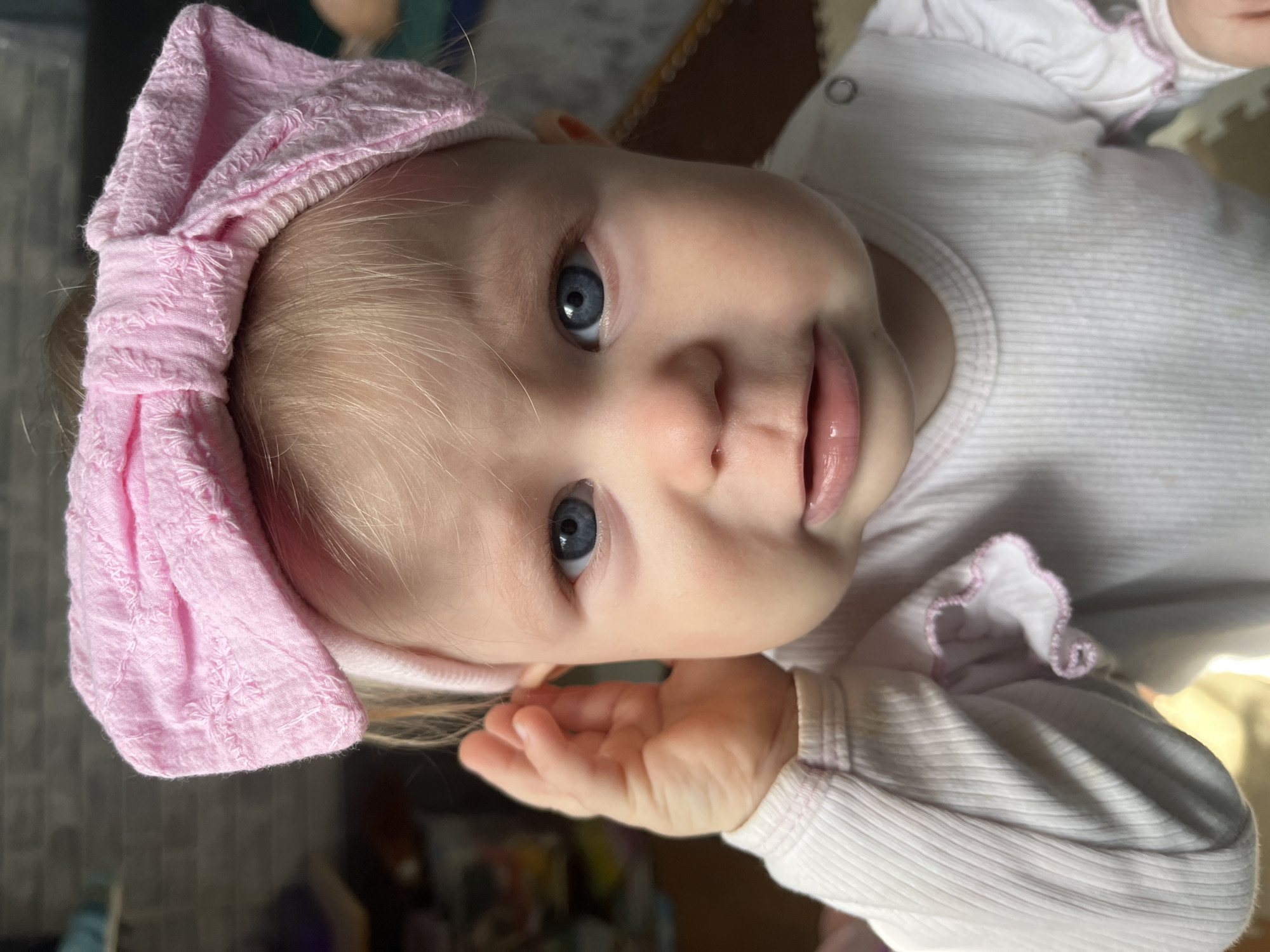 An overhead shot captures a baby or toddler looking directly at the camera. The subject has fair skin, blonde hair peeking out from under a pink bow headband, and striking blue eyes. The baby's mouth is slightly open, revealing pink lips. They are wearing a light-colored, possibly white or pale pink, ribbed long-sleeved top with a subtle pink trim around the neck and cuffs. A silver snap is visible on the shoulder of the top. One small hand is partially visible, curled near the baby's chin. The image is taken from an oblique angle, with the baby's head tilted towards the left side of the frame. The background is softly blurred, indicating an indoor setting, likely a home, possibly on a bed or a soft playmat. The lighting suggests a well-lit interior. No specific location cues are visible within the image to identify Vesyoloye, Russia. There is no visible text. The baby appears calm and curious, engaging with the camera.