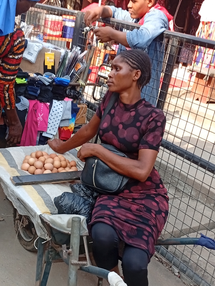 A woman sits on the edge of a cart, wearing a burgundy polka dot dress and black leggings. A black purse rests on her lap. The cart displays a pile of brown eggs on a striped cloth and an assortment of socks and other items. A man in a denim jacket stands behind the cart, and part of another person wearing patterned sleeves is visible on the left. A metal fence surrounds the scene.