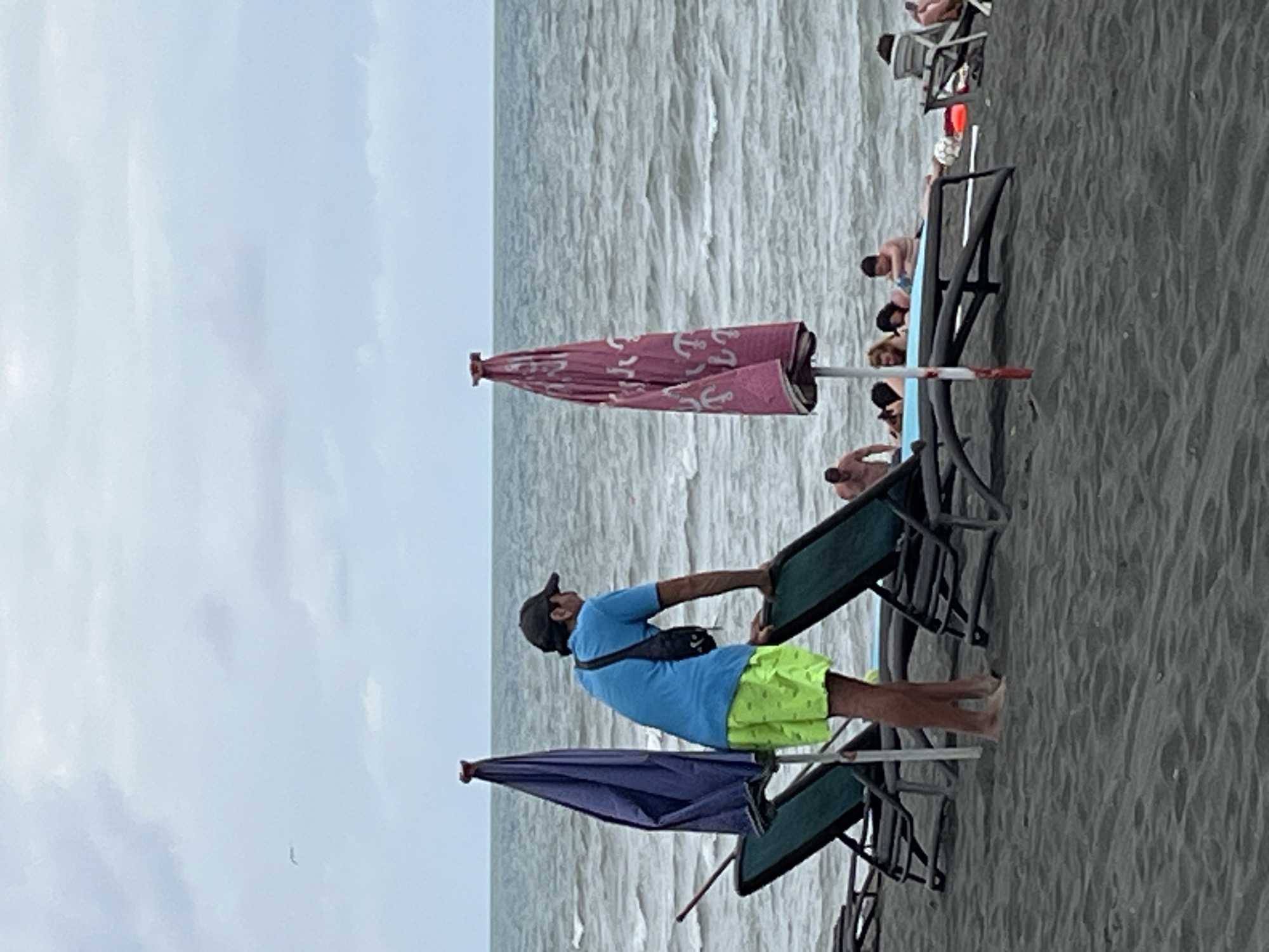 The image shows a sandy beach in Ureki, Georgia, with the Black Sea in the background. The sky is overcast. In the foreground, a man wearing a blue t-shirt and bright green shorts stands leaning on a beach chair. Two beach umbrellas are visible, one dark blue and the other pink with a repeating anchor pattern. In the middle ground, several people are relaxing on beach chairs or lying on the sand near the water. One person is partially visible in the lower left corner of the frame. The overall scene depicts a typical day at the beach.