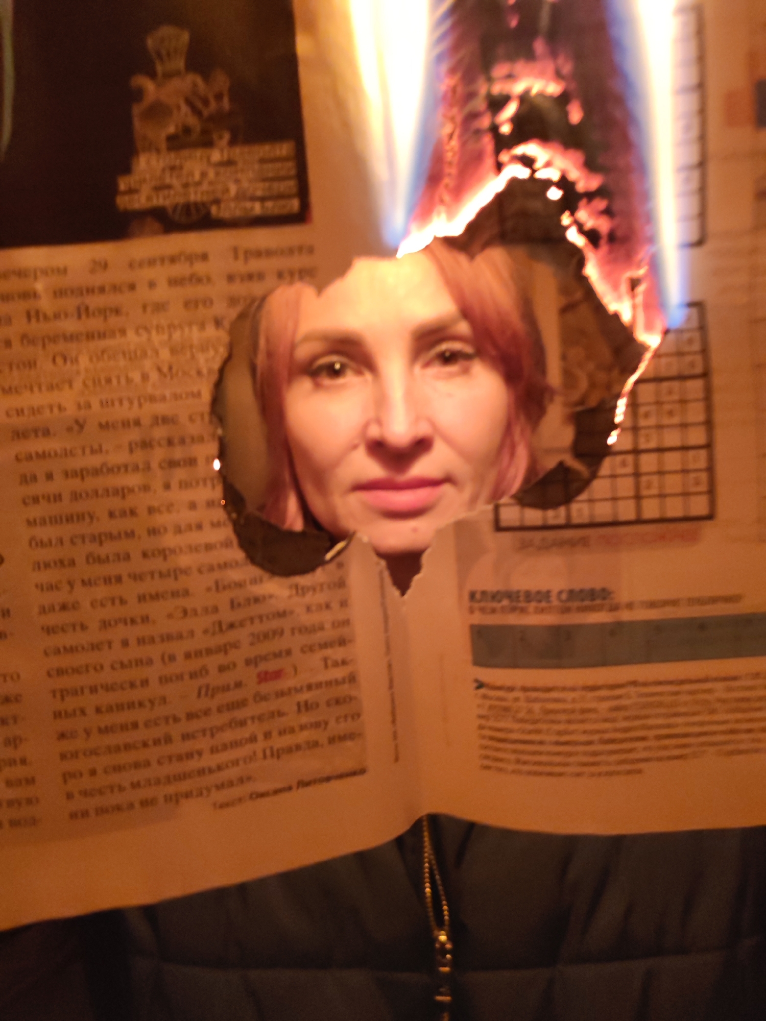 A woman's face is seen through a hole burned in a page of a newspaper. The edges of the hole are still aflame. The newspaper page is held up against a dark jacket. The article in the newspaper is in Russian. A crossword puzzle and the heading "KEY WORD" are visible on the right side of the paper.