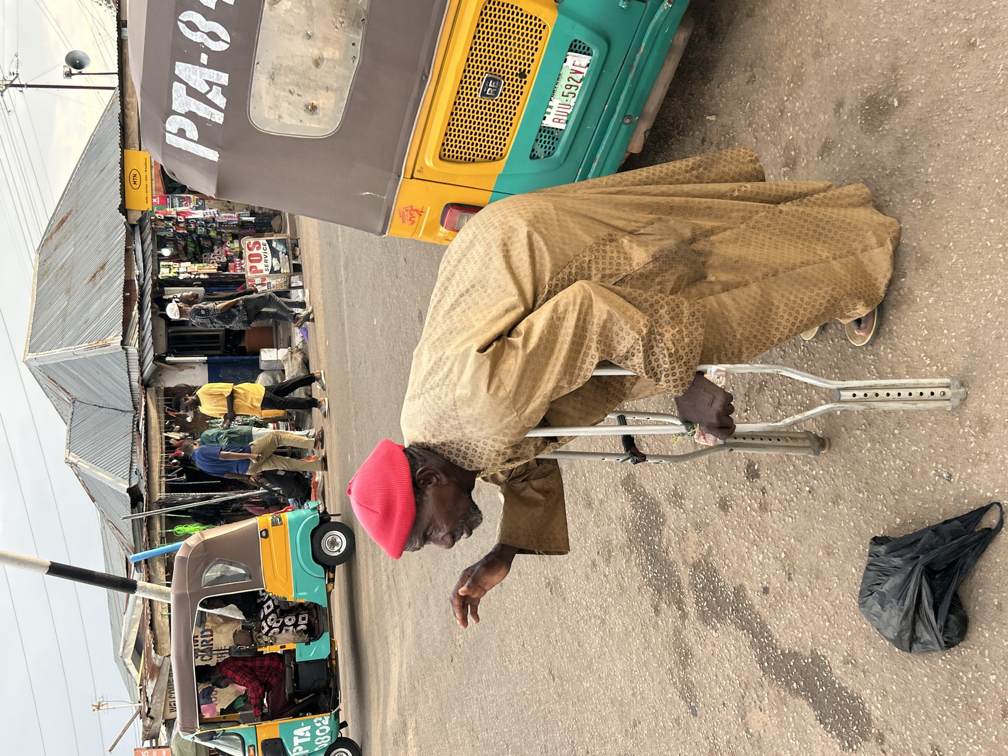 This image, captured in Jos, Nigeria, depicts a bustling street scene, likely a market or commercial area, under an overcast or hazy daytime sky.
In the foreground, an elderly man with dark skin, wearing a light brown patterned tunic (agbada-style) and a bright red cap, leans on a pair of metal crutches. His gaze is directed downwards and slightly to his right, and his right hand is partially extended with the palm open. A crumpled black plastic bag lies on the asphalt ground near his crutches.
Behind the man, two tricycles, commonly known as Keke Napep, are visible. The closest one is distinctive with its yellow, green, and brown coloring, displaying a license plate "BUU-592VE" and text "PTA-8..." on its side. Another similar yellow and green tricycle, bearing the text "PTA-0802," is partially visible further left in the background.
The background features a row of simple shop fronts, constructed with corrugated metal roofs and walls. These shops display various goods, and several other individuals are visible standing or moving near them, engaged in daily market activities. Prominent signage includes a yellow "MTN" logo on one shop, and another sign advertising "POS SERVICE" and "CARD" along with the word "WELCOME." A utility pole stands on the left. The ground is paved asphalt, showing signs of wear.