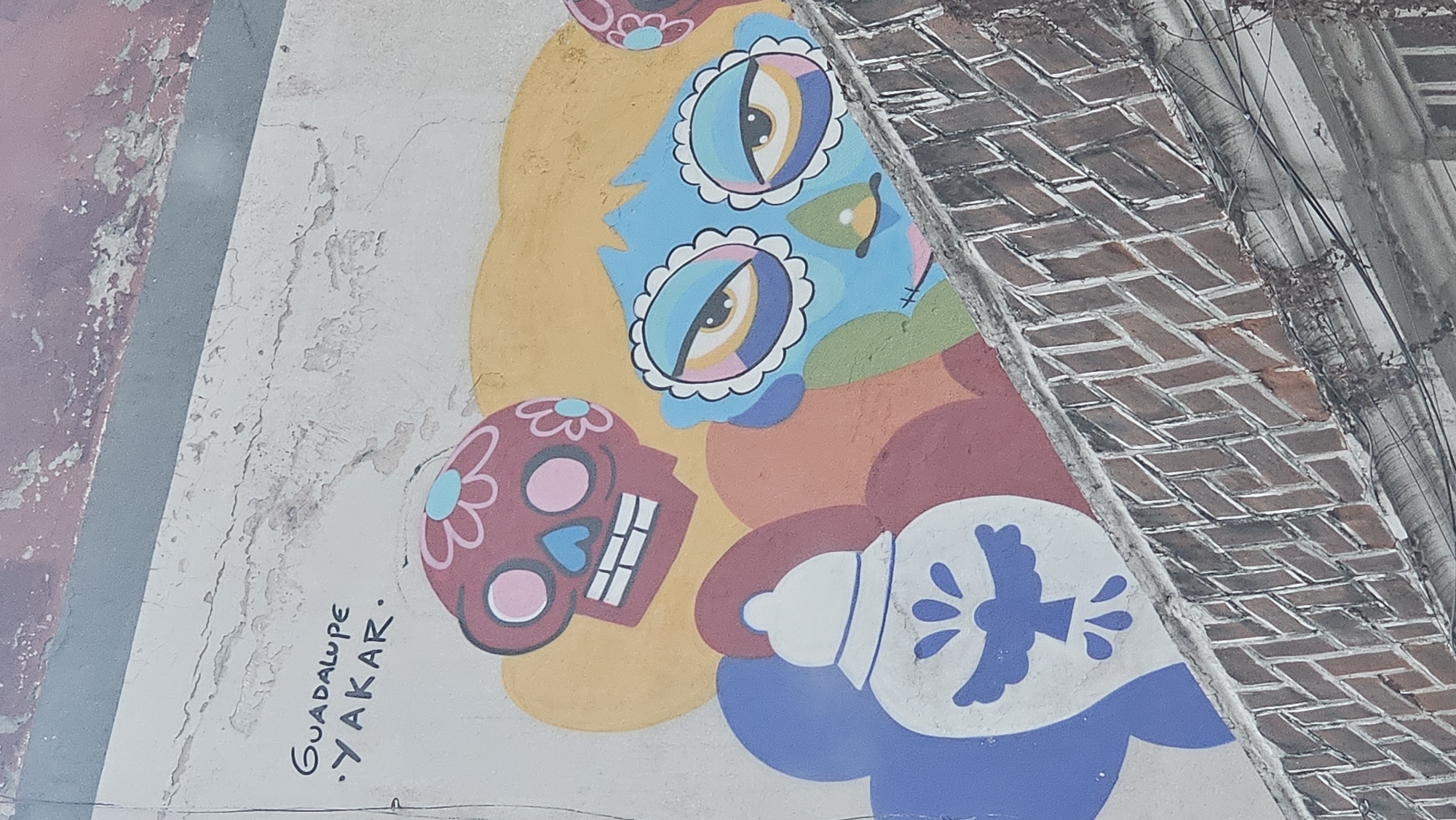 A colorful mural is displayed on a textured, light-colored wall in an urban setting, likely in Puebla, Mexico. The image is rotated, showing the mural on its side. The wall exhibits signs of age, with peeling paint and cracks. To the right, a diagonal section of a red brick wall is visible, accompanied by some wires and dried vegetation, indicating an outdoor location.
The mural features a large, stylized blue-faced figure with prominent, expressive eyes outlined with white frills, each pupil comprising concentric rings of blue, pink, yellow, and brown. A small green nose-like shape is present on the face, and a large yellow, cloud-like shape frames the figure's head, resembling hair or a halo. To the left and upper-left, two smaller, dark pink or maroon sugar skulls (calaveras) are depicted. The more prominent sugar skull has white teeth, pink eye sockets, a blue heart-shaped nose, and a light blue flower on its forehead.
Below the main figure, a white, bottle-shaped object with a blue dove motif, surrounded by stylized splatters, rests on a blue, cloud-like base. The background of the mural is filled with overlapping, organic shapes in vibrant colors such as yellow, orange, green, and red. The overall style blends traditional Mexican folk art elements with a contemporary, cartoonish aesthetic, creating a cheerful and culturally rich visual.
The artist's signature is clearly visible on the lower-left portion of the mural, reading "Guadalupe .YAKAR.". The scene captures a static artistic display, rich in color and cultural symbolism, characteristic of street art in Mexican cities. The lighting suggests it is daytime.