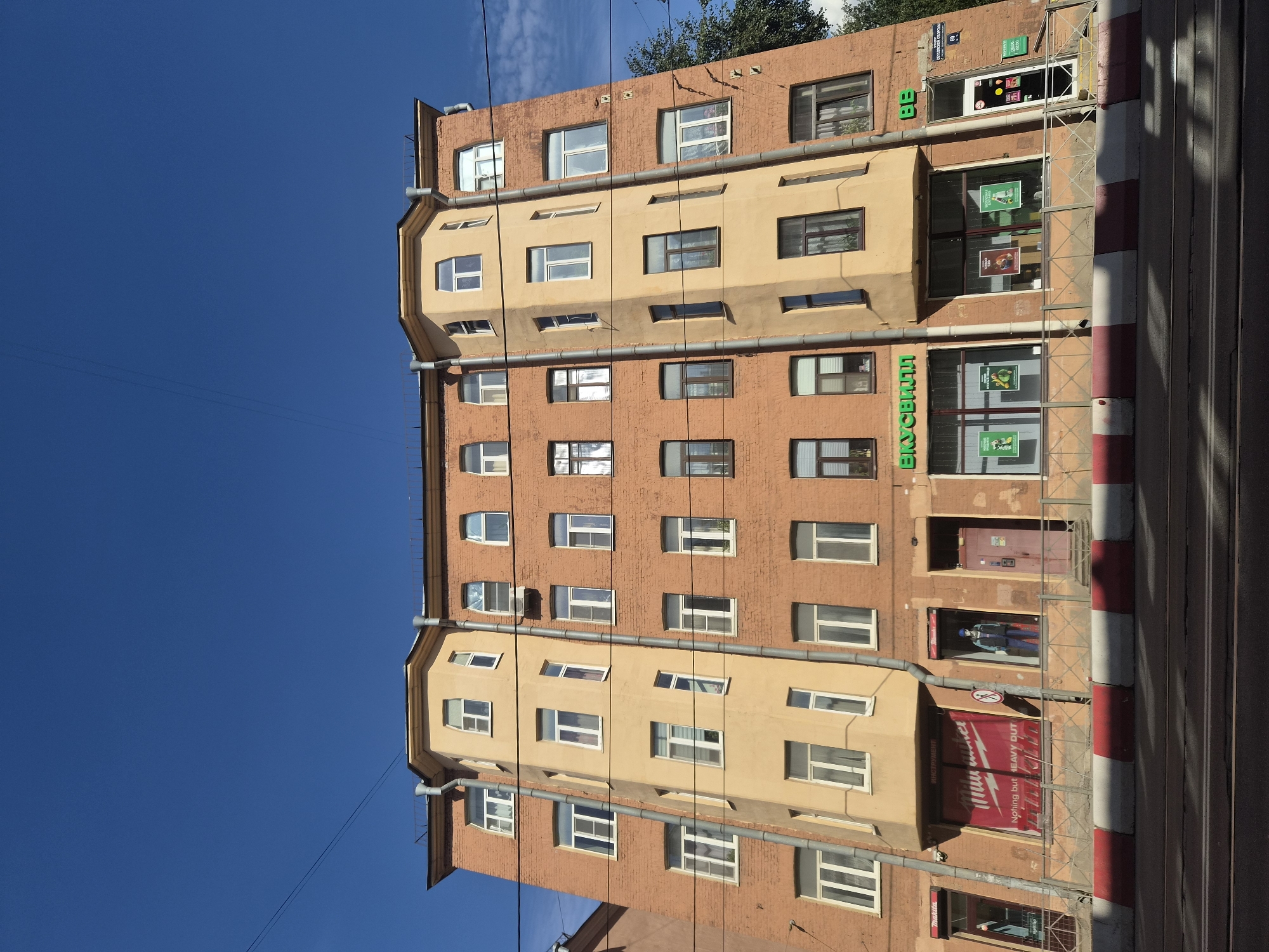 The image depicts a multi-story brick building on a sunny day in Saint Petersburg, Russia. The building has an orange-brick facade with some sections painted a light yellow. It features numerous windows of various sizes, many with white frames. The architectural style appears to be of the early 20th century, characterized by its solid construction and symmetrical window placement.
On the ground floor, there are several commercial establishments. One storefront displays a large banner for "Milwaukee" tools with the slogan "Nothing but HEAVY DUTY". Another section features a store with green signage that reads "ВКУСВИЛЛ" (VkusVill), a Russian supermarket chain, displaying posters in its windows. Adjacent to VkusVill is another store with green signage "BB". There is a red door leading into one of the buildings.
A metal fence runs along the sidewalk in front of the ground floor shops. Across the street, a tram track is visible, along with a red and white striped traffic barrier. Power lines crisscross the clear blue sky above the building, and a hint of green foliage from a tree can be seen on the right side of the image. The overall impression is of a typical urban street scene in a historic European city. The building is located on a street in Saint Petersburg.