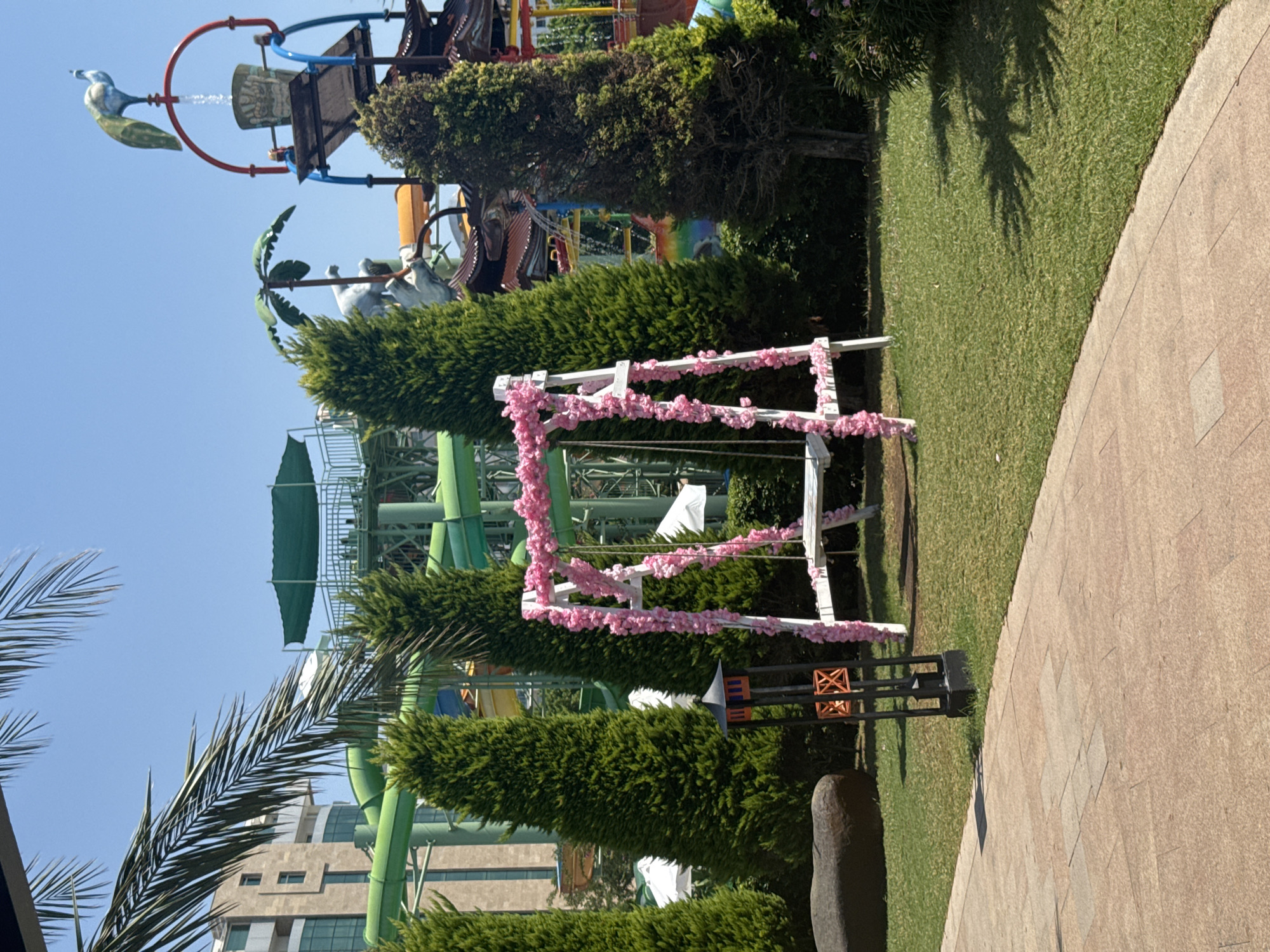 The image captures an outdoor scene at a water park, likely in Aksu, Turkey, on a sunny day. In the foreground, a pink-flowered white swing set is positioned on a lush green lawn. To the left of the swing, a large dark rock and a black decorative lantern with orange accents are visible. Behind the swing and to the right, tall evergreen trees provide a backdrop. Further in the background, several water slides, including green and yellow ones, are prominent, along with a building structure and a water feature with a peacock statue at its apex. The sky is clear and blue.