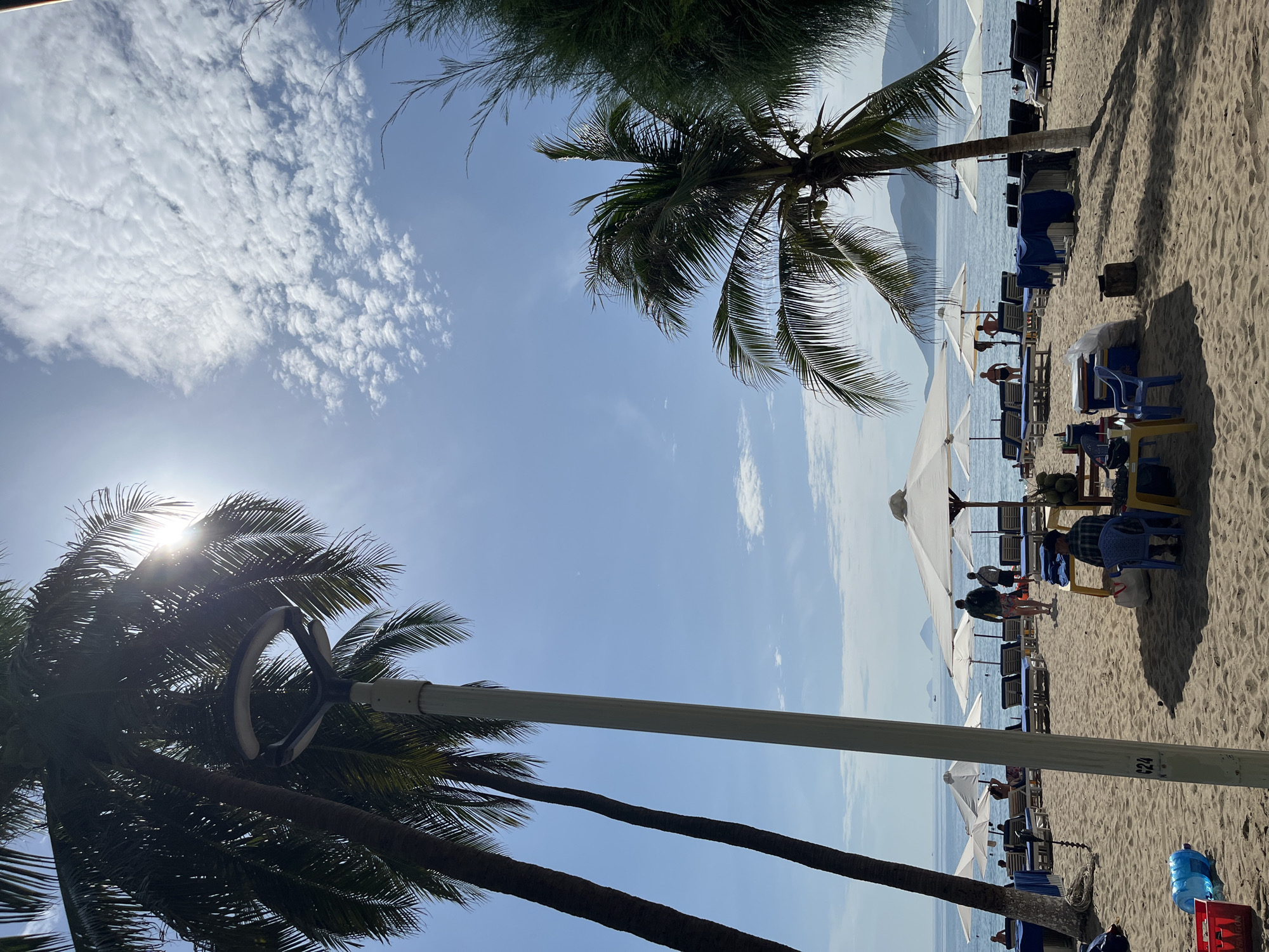 This image depicts a beach scene in Nha Trang, Vietnam, likely during daylight hours. The foreground is dominated by sand, with several white beach umbrellas casting shadows. Beach chairs, some covered with blue tarpaulins, are arranged in rows. In the middle ground, several people are visible, some walking and others seated at tables. To the left, a tall light post stands next to a palm tree. Coconut trees are also prominent throughout the scene, with their fronds framing the sky. The background features the calm blue ocean, with distant islands or hills visible on the horizon. The sky is clear blue with scattered white clouds, and the sun is shining brightly, with its rays partially obscured by palm fronds, creating a lens flare effect. The overall impression is of a typical beach resort atmosphere.