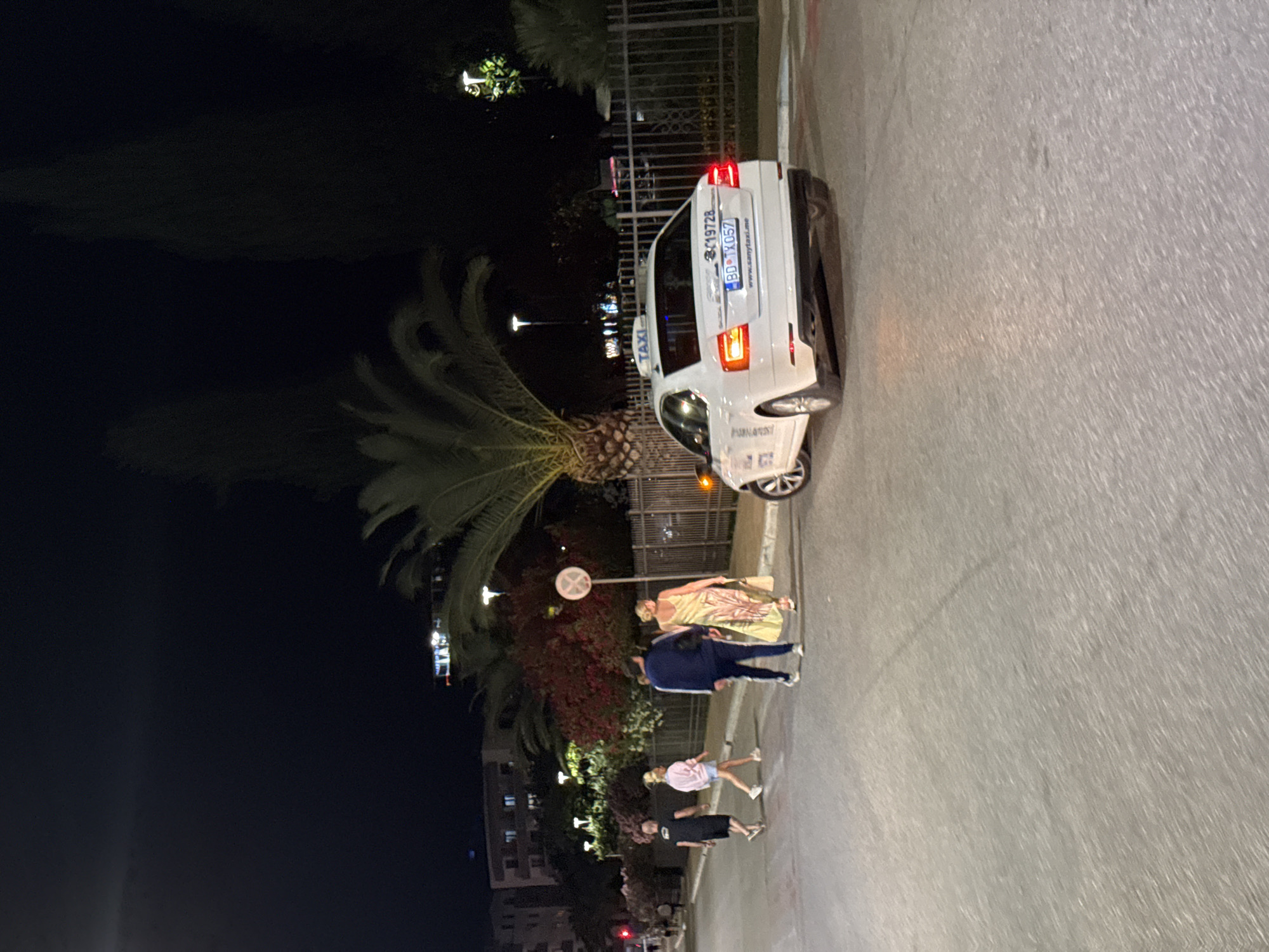 The image depicts a street scene at night in Budva, Montenegro, focusing on a white Skoda taxi. Several individuals are visible, some crossing the street and others walking along the sidewalk. The taxi has a visible license plate and a website address printed on the rear bumper. The background includes streetlights, palm trees, and buildings, suggesting an urban environment. The overall lighting is dim, characteristic of nighttime.