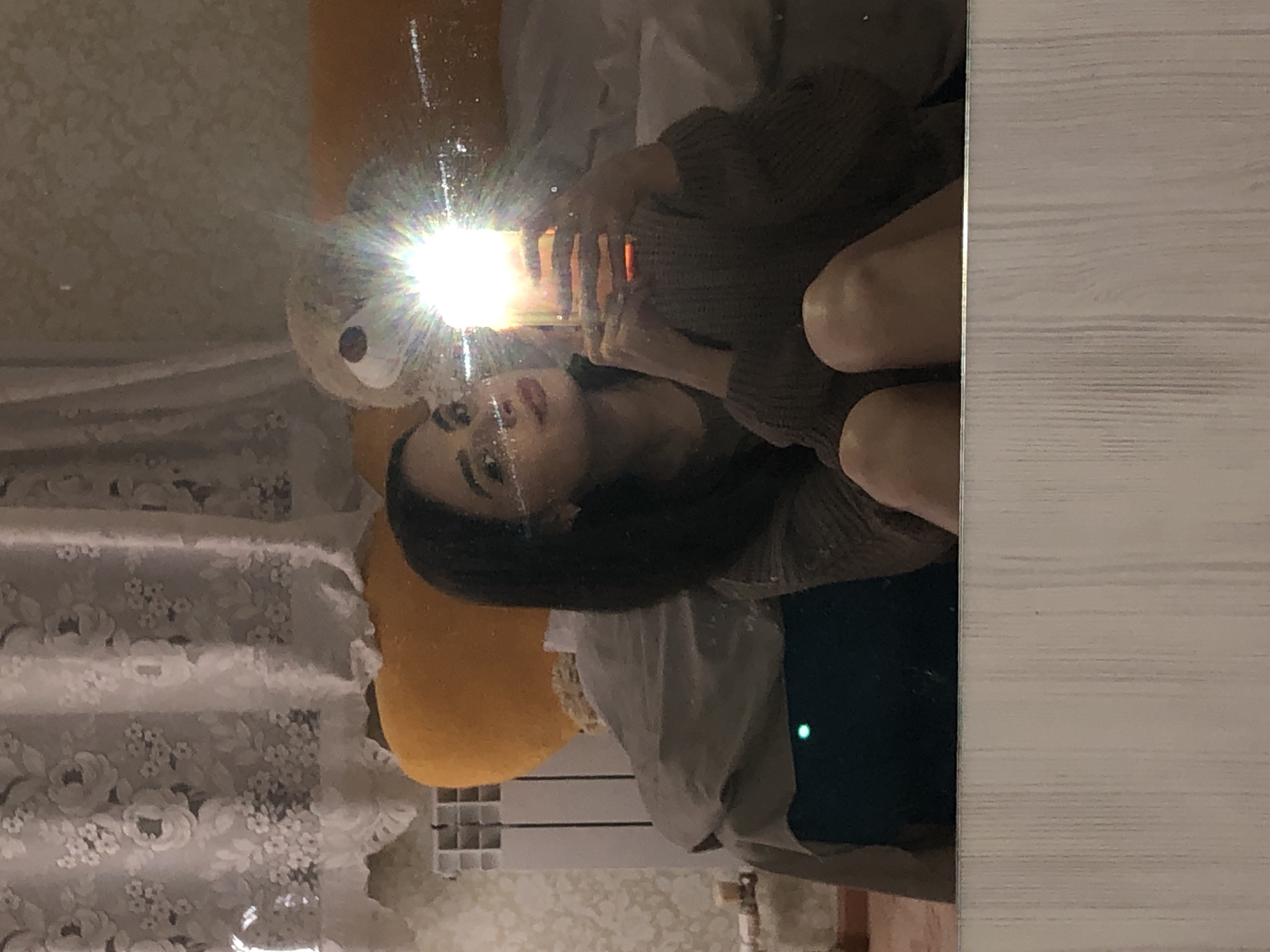 A mirror selfie captures a woman in a brown sweater holding a lit smartphone. The light reflects off the mirror. Visible in the background are a stuffed bear, decorative curtains, a radiator, and floral wallpaper.