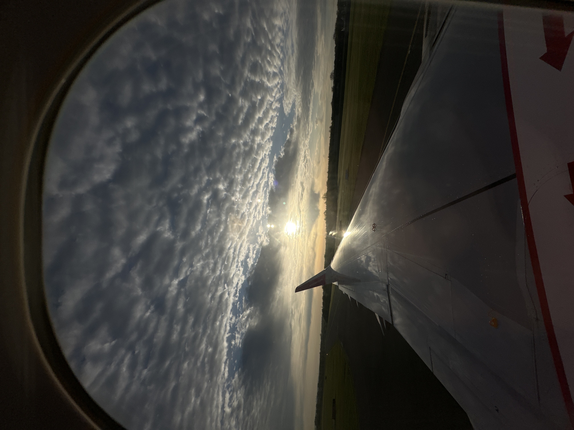 The image shows a view from an airplane window, with the plane's wing visible. The sun is shining brightly, creating a lens flare, and partially obscured by patchy clouds. Below, the ground is visible, seemingly a rural or semi-rural landscape with fields, possibly near Holzwickede, Germany, based on the Wizz Air livery on the winglet indicating the plane's location and route. The image is taken during sunrise or sunset, given the position of the sun.