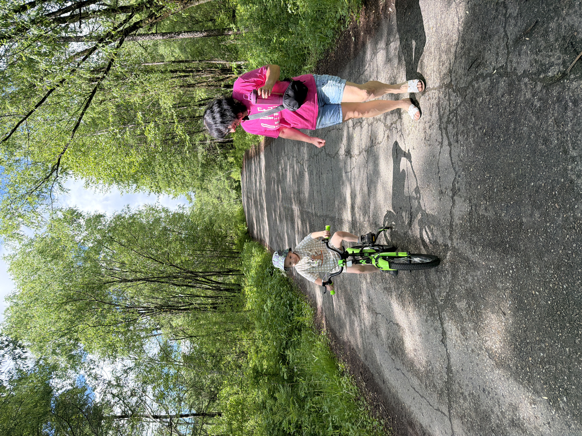 A full-shot photograph captures a paved path winding through a lush, tree-lined forest. The path is cracked and worn, with dappled sunlight illuminating patches of the asphalt. On the left side of the path, a young child, wearing a light-colored t-shirt with a graphic print and a bucket hat, rides a bright green bicycle with training wheels. The child is looking ahead, appearing to be in motion. On the right side of the path, a woman with dark hair, wearing a pink t-shirt and denim shorts, walks towards the child. She is holding a pink water bottle in her right hand and has a black fanny pack slung across her body. Her gaze is directed towards the child. The surrounding environment is dense with green foliage and tall trees, suggesting a natural, possibly rural or park-like setting. The sky above is a bright blue with scattered clouds. The location is identified as Kul'dur, Russia, likely a natural area or park within or near the city.