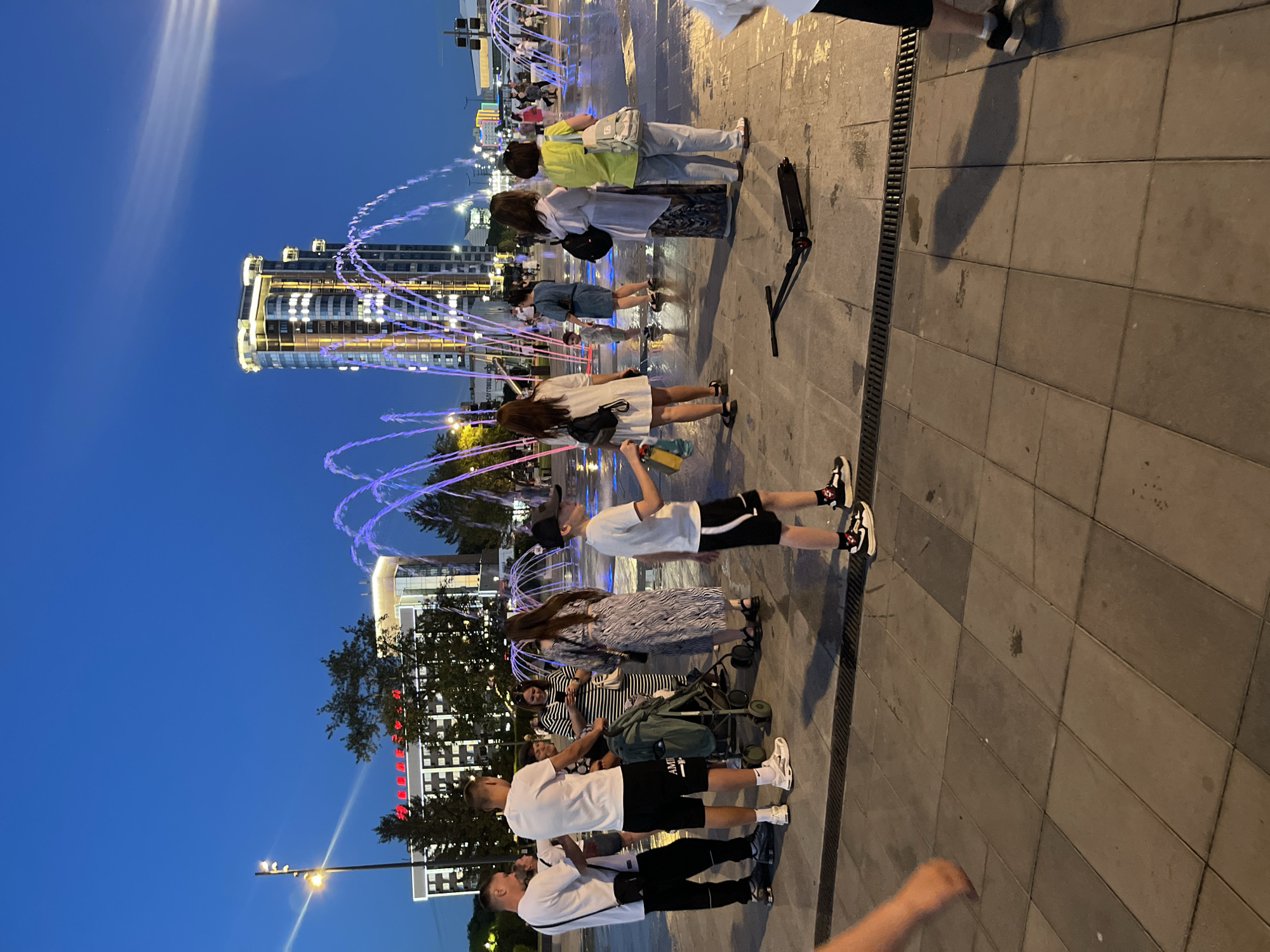 The image depicts a group of people gathered in an outdoor plaza at dusk, illuminated by artificial lights and a fountain. The setting appears to be a modern urban environment with tall buildings visible in the background. A fountain with illuminated water jets creates a dynamic visual element in the scene. Several individuals are standing on a paved surface, some are looking at the fountain, while others seem to be interacting with each other. One person is holding a scooter. The time of day is suggested by the deep blue sky and the artificial lighting, indicating it is evening or twilight. The atmosphere seems relaxed and casual, with people enjoying the public space. There is no visible text that can be deciphered from the image. The overall scene suggests a common urban leisure activity, such as socializing or sightseeing.
