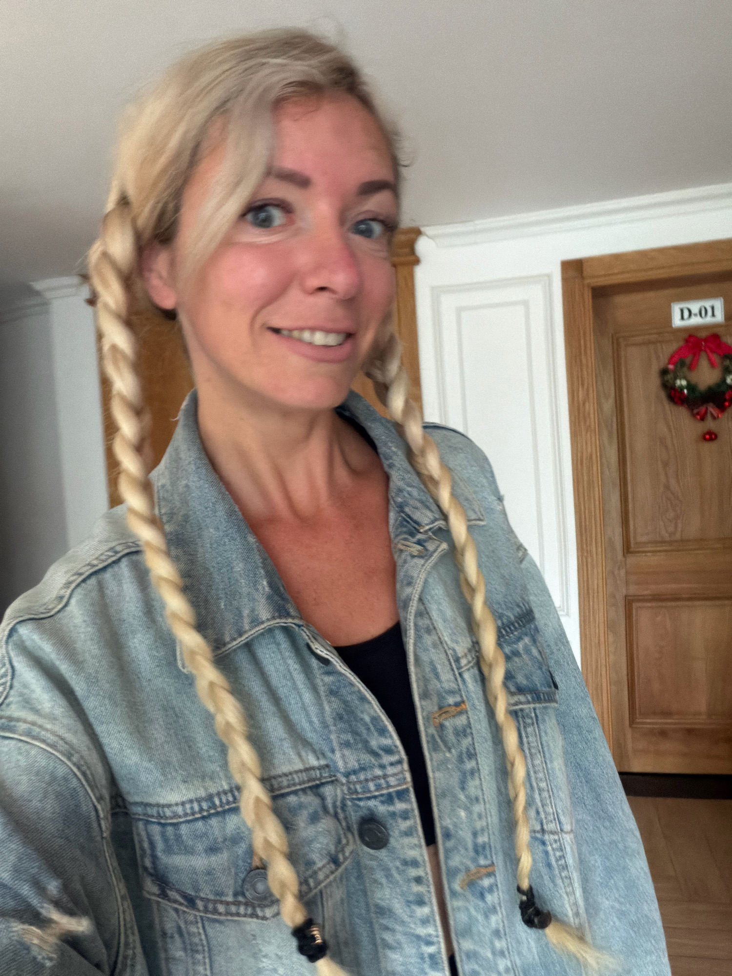 A blonde woman with her hair in two braids, wearing a denim jacket and a black top, smiles at the camera. She is standing indoors, in front of a wooden door with a wreath and a sign that reads "D-01". The background also shows white paneling on the wall and a glimpse of a ceiling. The overall impression is casual and possibly festive. The image does not provide any information about the location being Xuân Trường - Đà Lạt, Vietnam.