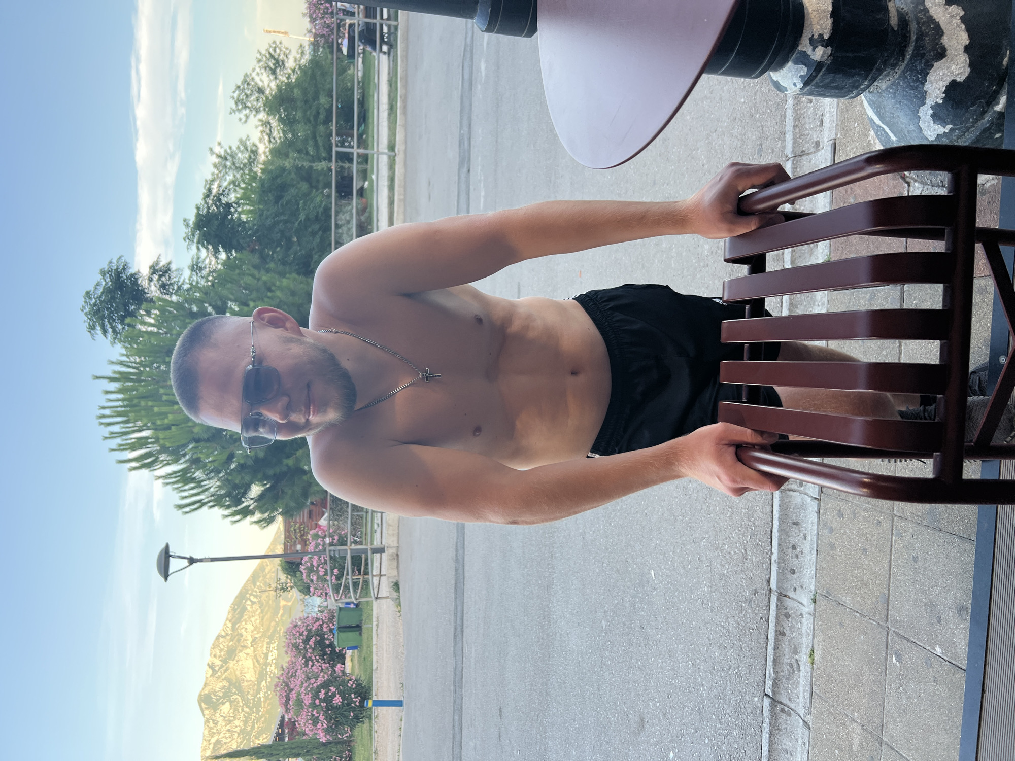 The image depicts a shirtless young man with short hair and sunglasses, wearing black shorts, standing outdoors. He is leaning on the back of a dark red chair, with his hands gripping its frame. His left arm is bent, resting on the chair, while his right arm is slightly extended. The man has a silver necklace with a cross pendant.
The background reveals a street or pathway with a paved surface and a concrete curb. Beyond that, there's a landscaped area with trees, including tall evergreen trees and flowering bushes with pink blossoms. In the distance, a mountain range is visible under a partly cloudy sky. A street lamp stands to the left of the frame. A dark round table with a textured base is partially visible on the right. The scene suggests an outdoor seating area, possibly at a cafe or restaurant in Budva, Montenegro, given the mountainous landscape and Mediterranean vegetation.
