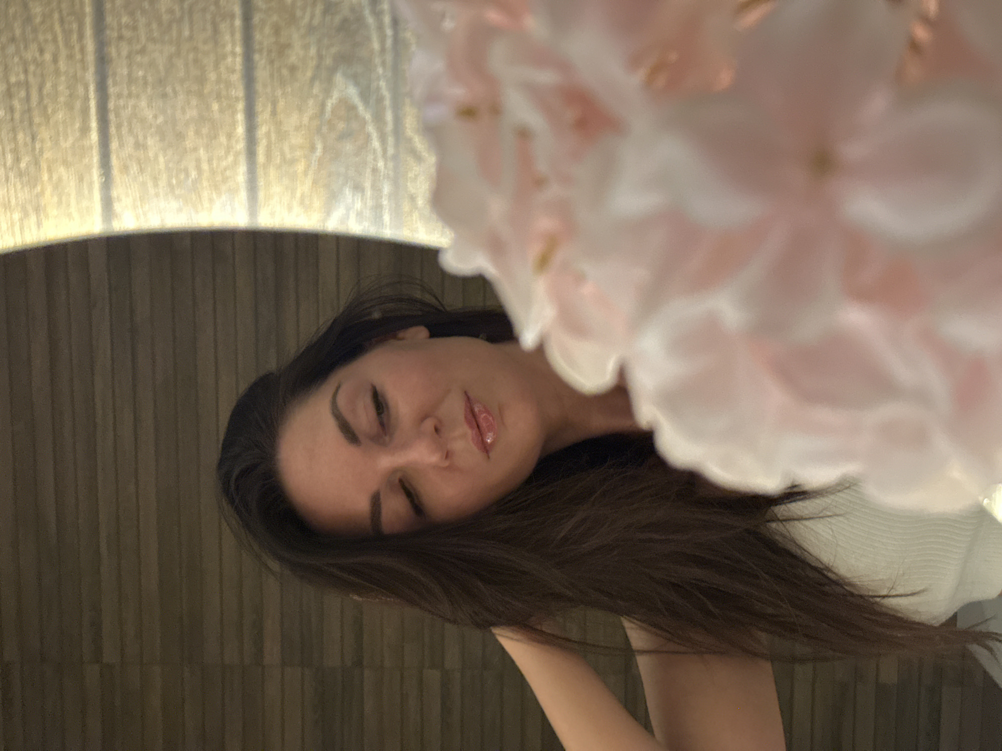A medium shot presents a woman with dark hair partially obscured by blurred pink and white flowers in the foreground. The woman’s face is visible, displaying closed eyes, dark eyebrows, and light pink lips. The background includes a wall with vertical wood paneling. A curved, lit surface is above the woman's head. The location in the city of Chaikovsky, Russia, cannot be determined from the image.