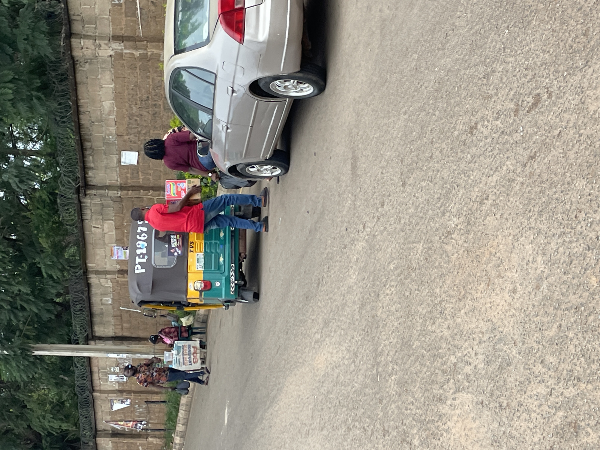A medium shot captures a street scene in Jos, Nigeria, dominated by a silver sedan and a colorful auto-rickshaw parked on a paved road. The auto-rickshaw, predominantly yellow and green, displays the license plate "PT-19679". Behind the vehicles, a light brown brick wall topped with barbed wire runs along the background, partially obscured by lush green trees. Several posters are attached to the wall. In the foreground, the road surface is a textured gray asphalt.
Two men and two women are present. One man, wearing a red t-shirt and blue jeans, is visible from the chest down, facing towards the right and holding a box. Another man, in a patterned short-sleeved shirt and dark pants, walks towards the left, carrying a black bag. Two women are near the auto-rickshaw. One, wearing a maroon shirt and dark pants, is near the silver car, and the other, in a pink top, is seated behind a small table on the left side of the road. The table has items on it, including what appears to be a POS machine and food items. The scene suggests a roadside commercial activity.