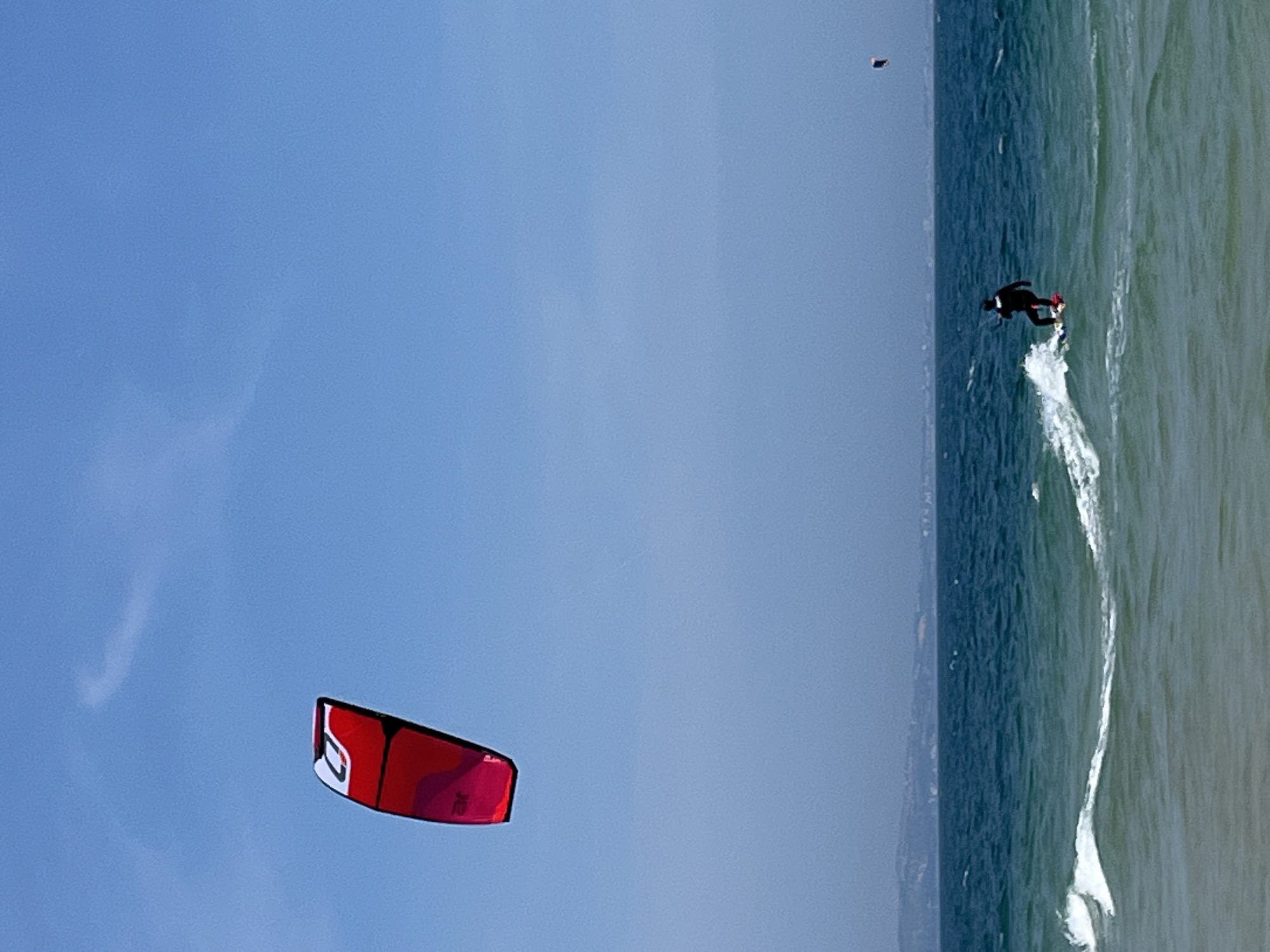 The image captures a bright, clear day with a vast expanse of blue sky meeting the deep blue ocean. In the foreground, the turquoise water shows gentle waves breaking. A lone figure, dressed in a black wetsuit, is kitesurfing on a red and white board, creating a white splash as they glide across the water. To the left of the frame, a large, red kite with white accents is visible, suspended high in the sky. Further in the distance, a faint coastline with hills is discernible under a hazy sky. Another smaller black and red kite is visible in the far right of the image. The scene appears to be a coastal area, possibly a beach or bay. The specific location in Phu Thuy Ward, Vietnam, is not visually identifiable from the image itself.