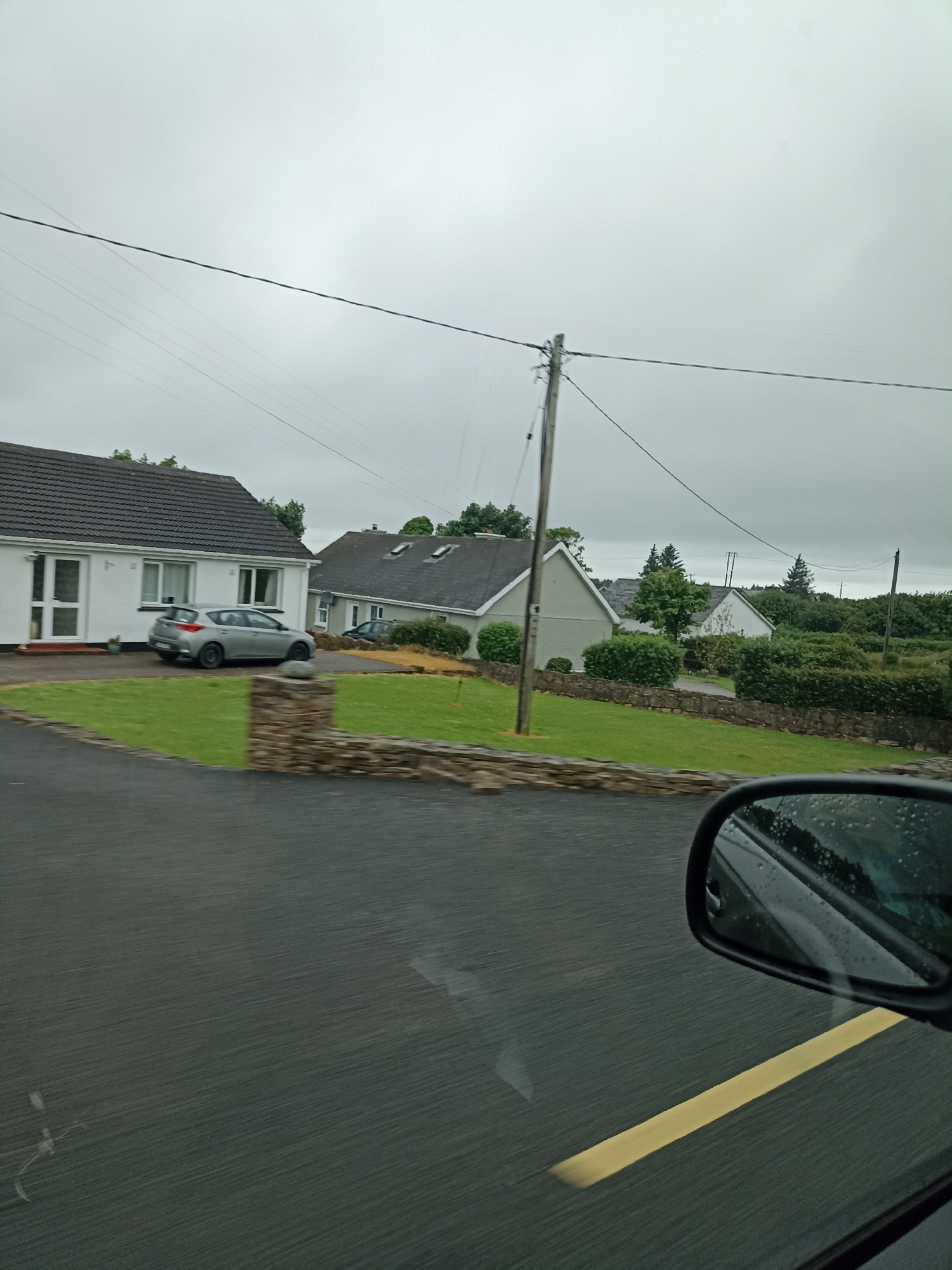 The image captures a view from a car, looking out at a residential street in Dore, Ireland, under an overcast sky. In the foreground, the wet asphalt road is visible, with a distinct yellow line marking its edge. A car's side mirror, reflecting the grey sky and some trees, is positioned in the lower right corner.
Further into the scene, a neatly maintained lawn slopes gently upwards, bordered by a low, rough-hewn stone wall. Beyond the wall, several houses are visible. To the left, a white bungalow with a dark tiled roof features large windows and a front door. A silver hatchback car is parked in the driveway.
To the right of this first house, another bungalow with a similar dark tiled roof and dormer windows is situated. A line of mature green hedges runs along the front of this property and continues to the right, obscuring the bases of the houses behind them. In the background, more houses and dense greenery, including trees, are scattered across the landscape. Power lines and a wooden utility pole are present, crossing the sky. The overall impression is one of a quiet, suburban or semi-rural setting on a damp, cloudy day.