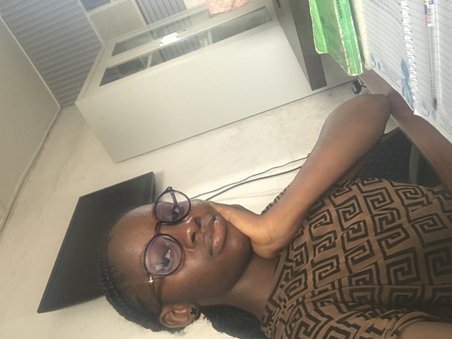 This image depicts an indoor scene with a young Black female, appearing to be in her late teens or early twenties, seated at a desk. She wears a brown short-sleeved top with a black geometric pattern, round tortoise-shell eyeglasses, and small black stud earrings. Her braided hair is visible. Her right hand is placed under her chin, and she maintains a neutral expression, looking directly at the camera. In the background, a white wall features a mounted flat-screen monitor on the left, with visible electrical wires. To the right, a tall white metal cabinet with glass doors is positioned beside a dark-colored desk. On the desk, multiple notebooks are visible, including one with a green spine partially labeled "NIGERIAN" and another open notebook with blue decorative elements and a transparent pen resting on it. The specific location within Ado, Nigeria is not discernible from the visual information provided in the image.