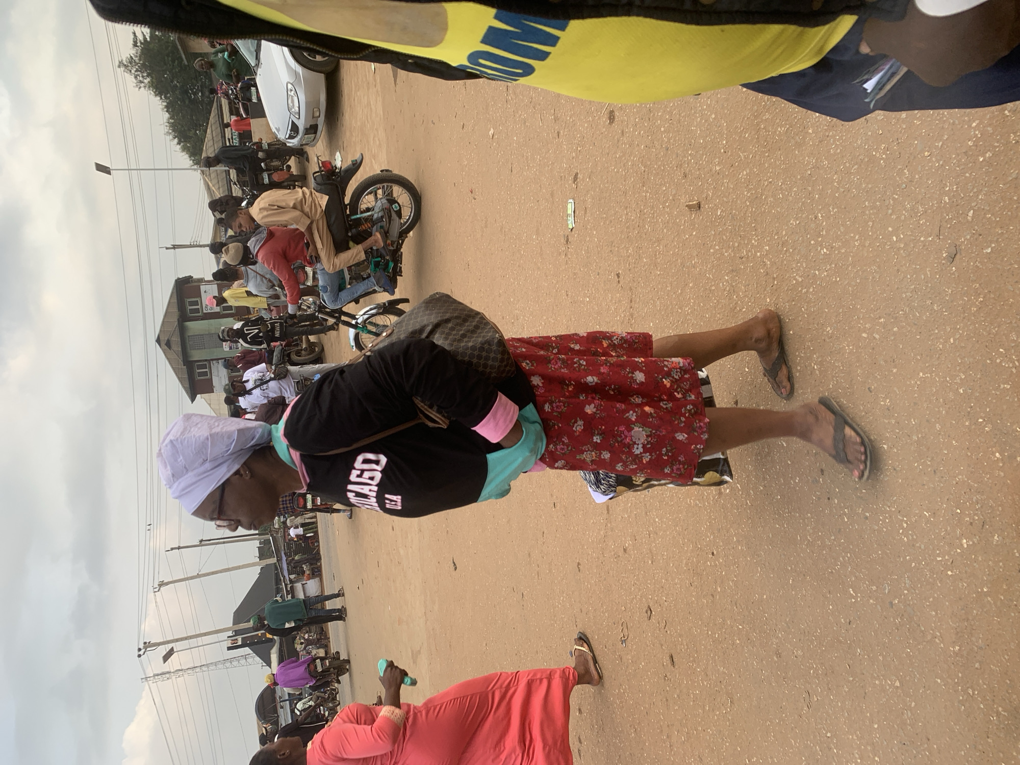 In Shagamu, Nigeria, a group of people are gathered on a dirt road under an overcast sky. A woman in a red dress and sandals walks away from the camera, holding a turquoise object. To her right, a woman wearing glasses, a white headscarf, a black sweatshirt with "CHICAGO" printed in white, and a red floral skirt stands facing away from the camera, holding a dark patterned handbag. She wears sandals. Further into the scene, a man in a light brown outfit rides a motorcycle with a passenger, both wearing sandals. Other individuals are seen in the background, some on motorcycles, and a silver car is parked. Buildings and utility poles are visible on either side of the road.