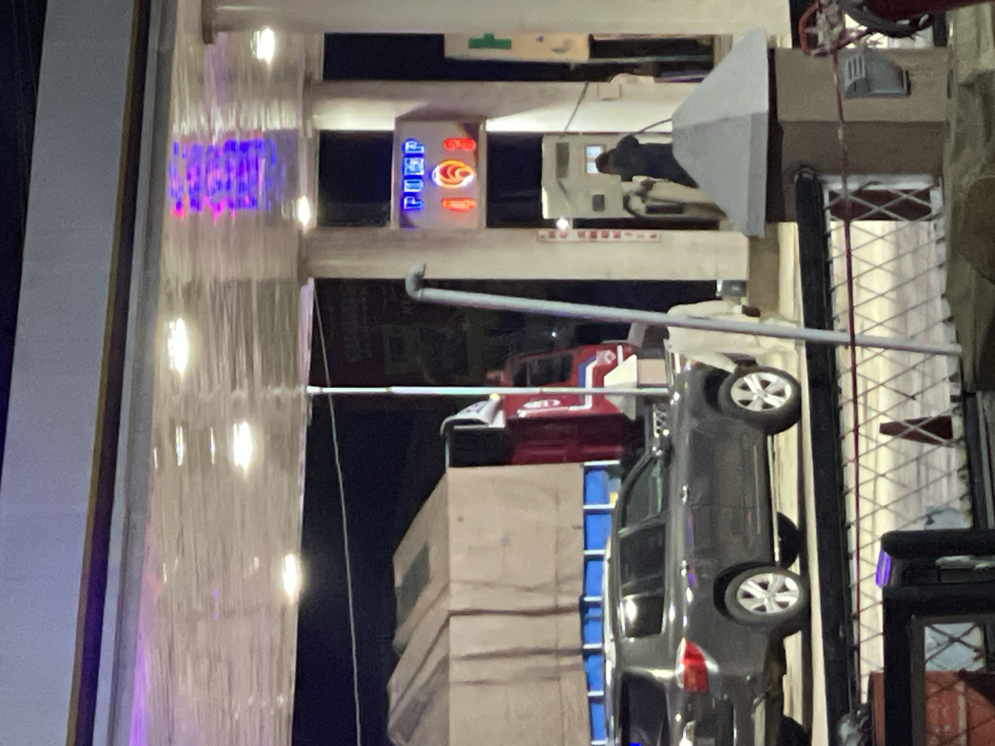 The image shows a gas station in Hayin Banki, Nigeria. A grey SUV is parked near a pump. A sign with the word "FUEL" is visible on the gas station structure. A red truck is also present. The station is lit by overhead lights. The environment appears to be urban with tall structures.