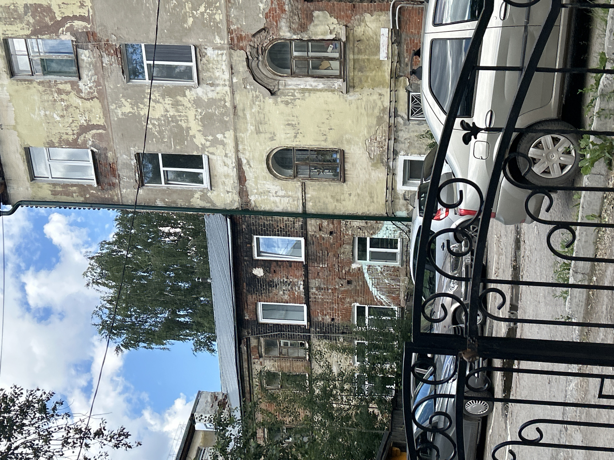 The image depicts a section of an old residential building in Perm, Russia, characterized by peeling paint, exposed brickwork, and various windows, some of which are modern replacements. The building's facade shows signs of age and weathering. In front of the building, several parked cars are visible, including what appears to be a silver sedan. A black wrought-iron fence or gate partially obstructs the view of the cars and the lower portion of the building. Trees and foliage surround the building, and the sky above is partly cloudy. The overall impression is of an urban environment with older architecture.