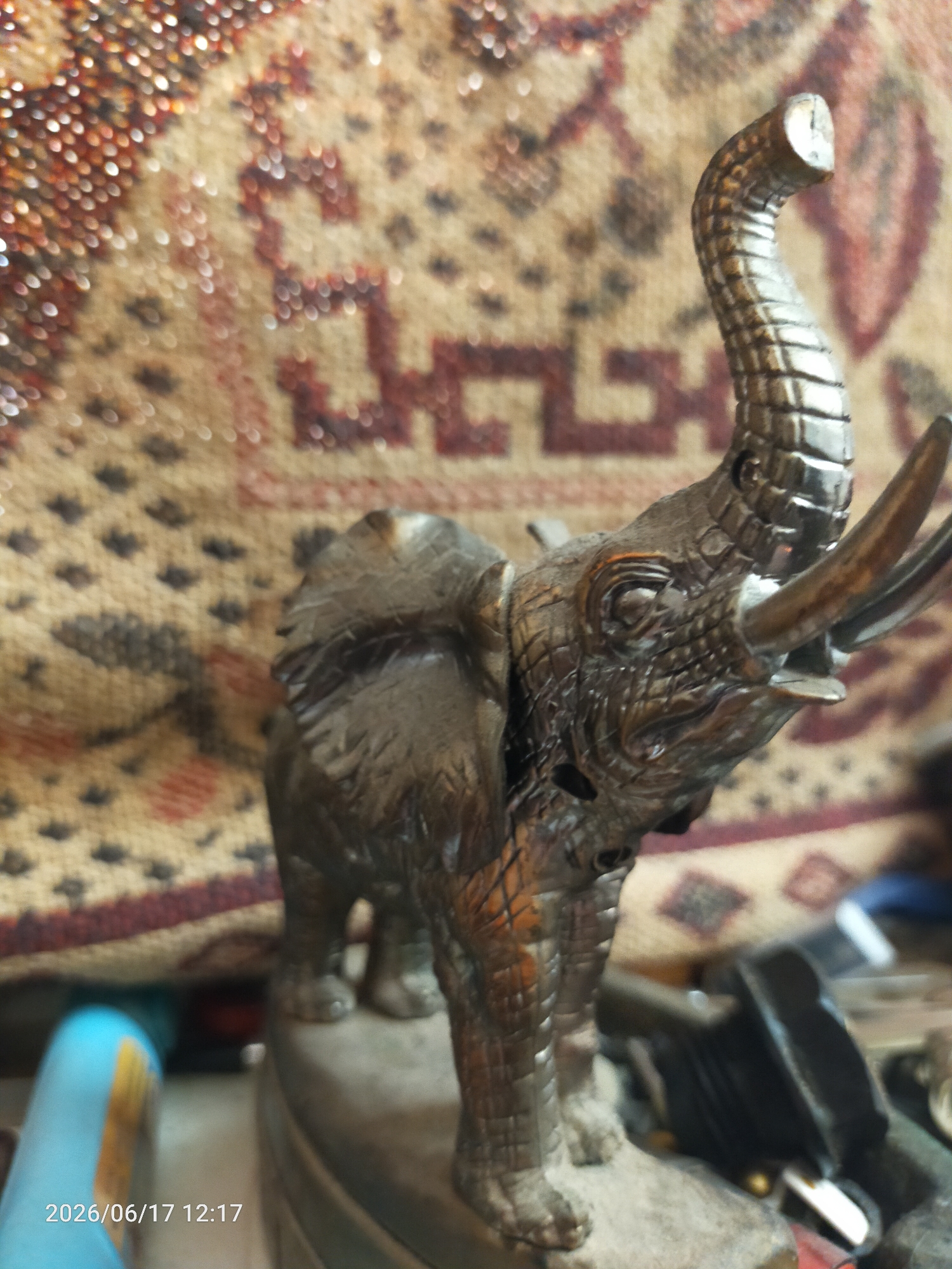 A detailed, low-angle, close-up shot features a metallic figurine of an elephant positioned on a rough, uneven surface. The elephant, rendered in a dark, reflective material, displays intricate detailing with textured skin and pronounced tusks. Its trunk is raised and curved. The background is a softly focused tapestry or rug with a geometric pattern in muted earthy tones, featuring reddish-brown and dark brown geometric shapes against a beige background. In the lower left corner, a blue cylindrical object with a yellow stripe is partially visible. The date stamp "2026/06/17 12:17" is overlaid on the image in the bottom left. The lighting creates highlights on the metallic surface of the elephant and gives a bokeh effect to the background. The overall scene appears to be an indoor setting, possibly a display or a shelf.