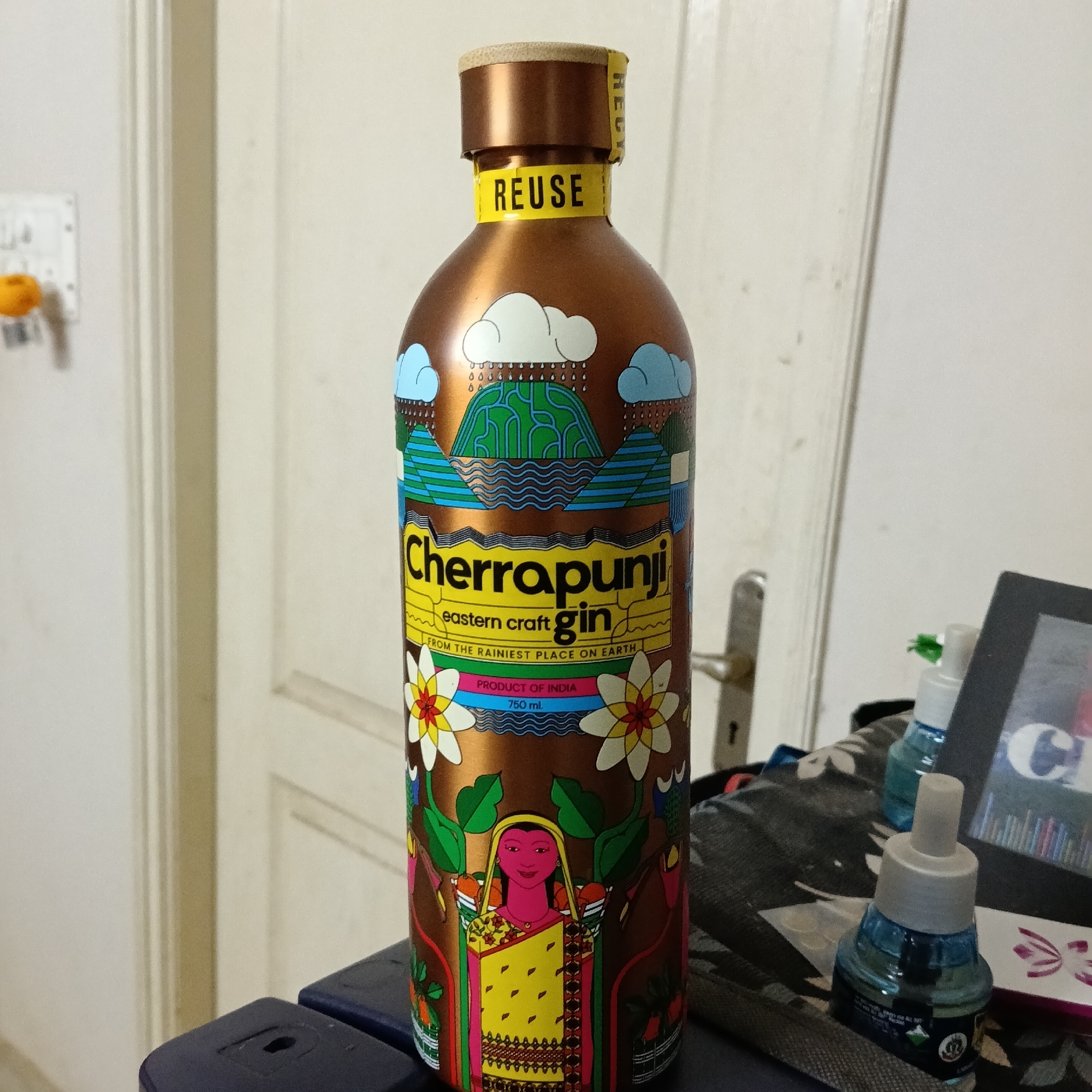 A copper-colored bottle of Cherrapunji eastern craft gin stands upright on a dark blue surface, possibly a table or a drawer. The bottle is decorated with an illustration depicting a rain scene with clouds and mountains, followed by the text "Cherrapunji eastern craft gin FROM THE RAINIEST PLACE ON EARTH". Below this, there are colorful floral designs and a depiction of a woman in traditional Indian attire. The bottle has a yellow label around its neck with the word "REUSE" printed in black. In the background, a light-colored wall with vertical paneling is visible, and a door handle can be seen to the right of the bottle. To the right of the bottle, on a patterned fabric surface, are two small blue plastic containers with white dispenser tops, resembling air fresheners or hand sanitizers. A dark rectangular object, possibly a tablet or e-reader, is also present on the fabric surface, displaying a colorful image. The overall scene suggests an indoor setting, likely a home environment. The image was taken in Bengaluru, India, as indicated by the user's request.