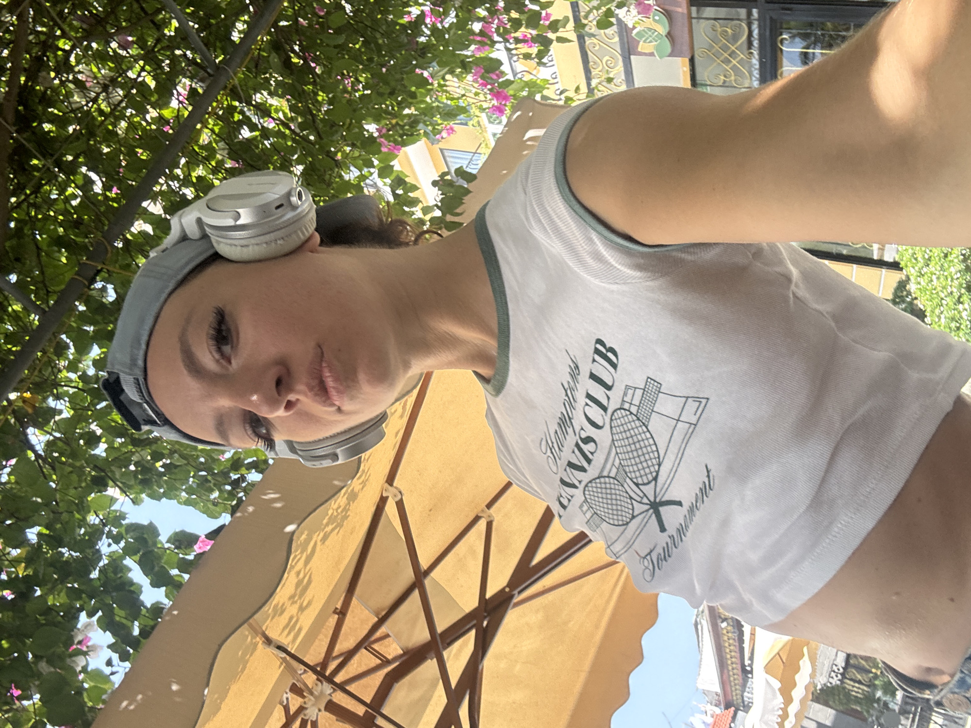 A waist-up selfie captures a fair-skinned woman wearing headphones, a grey athletic headband, and a white cropped shirt with a green trim around the neck. The shirt features a graphic depicting two tennis rackets and the text "Hamptons Tennis Club Tournament."
The shot is taken from a low angle, looking up at the woman. Overhead, a large, tan-colored umbrella provides shade. Beyond the umbrella, lush green trees with hints of pink flowers fill the upper left portion of the frame, while building facades appear in the lower right.
The scene is in Hội An Ward, Vietnam.