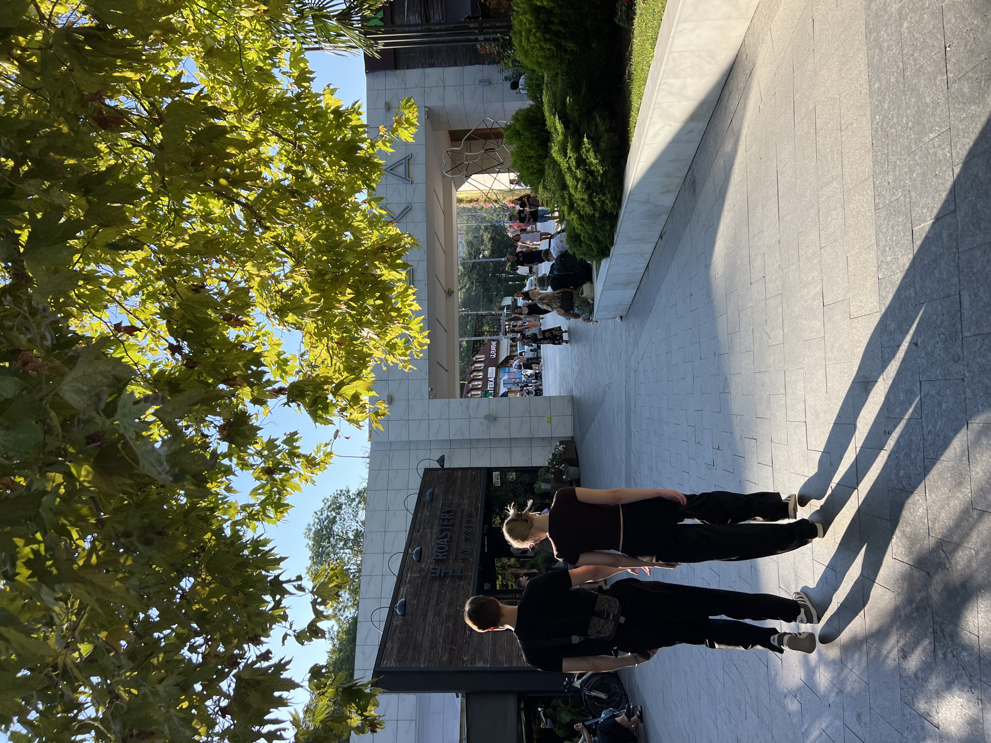 In Odesa, Ukraine, a young couple walks hand-in-hand on a sun-drenched pedestrian promenade. The man, on the left, wears a black t-shirt, black pants, and a crossbody bag. The woman, on the right, has on a dark brown cropped t-shirt and black pants. They are walking away from the camera, casting long shadows on the grey paved walkway.
To their left is a building with a wooden facade featuring the sign "ROASTERY" in white lettering. Above the building is a white structure with large letters, partially obscured by the foliage of a large tree with broad, green leaves, whose branches extend over the promenade, dappling the ground with sunlight and shadow.
Further down the promenade, other people are visible, some walking, others sitting. A white stone wall lined with manicured green bushes runs along the right side of the walkway. In the background, there is an arched entranceway and more buildings. The sky is clear and blue. The overall scene depicts a leisurely day in an urban outdoor space.