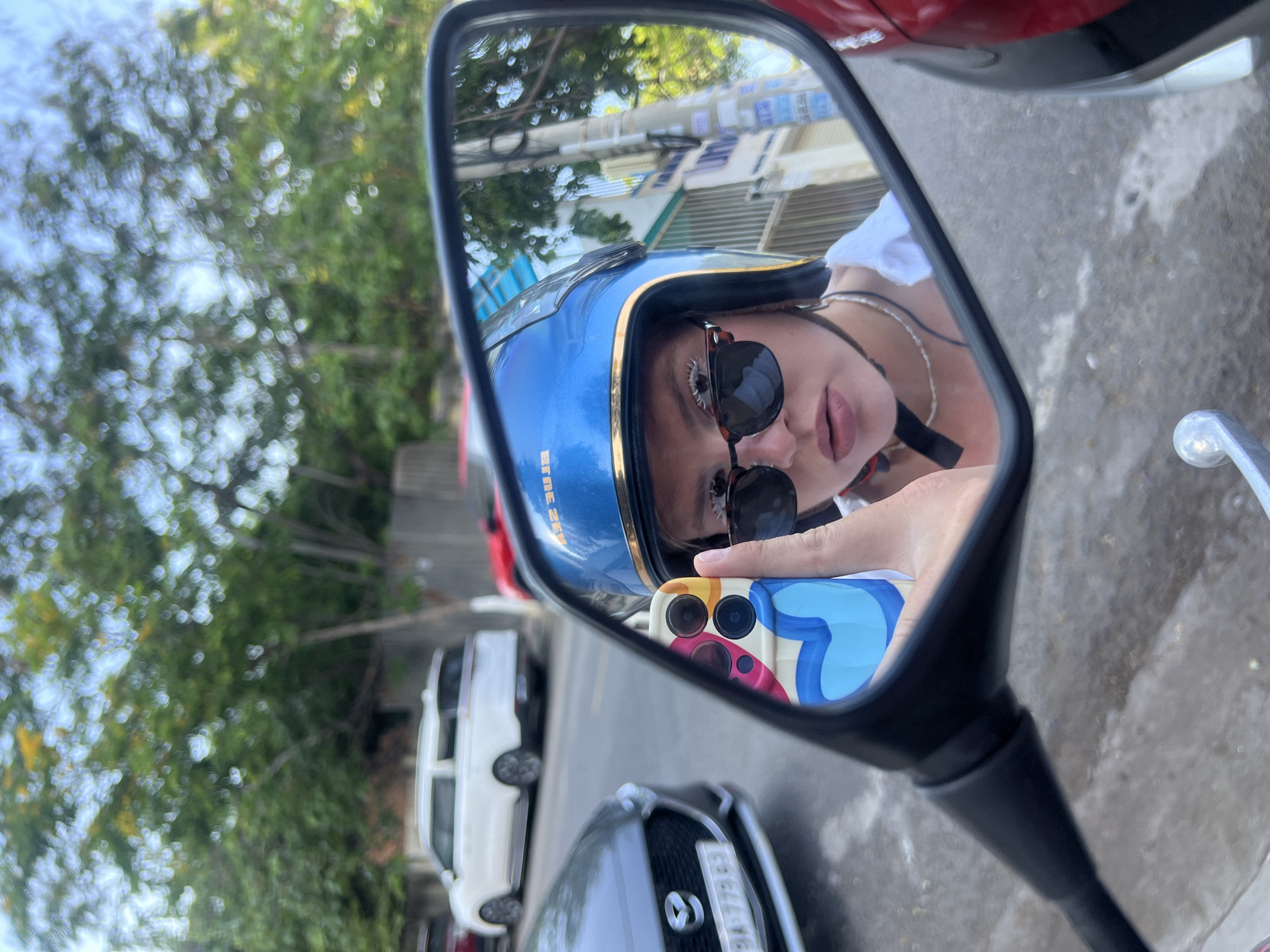 A selfie reflected in a motorcycle side mirror shows a person wearing a blue helmet and sunglasses. They are holding a phone with a colorful case. The reflection shows a street scene with trees, cars, and buildings. A white car and gray car are in the street. A Vietnamese license plate is visible. The location is Nam Nha Trang Ward, Vietnam.