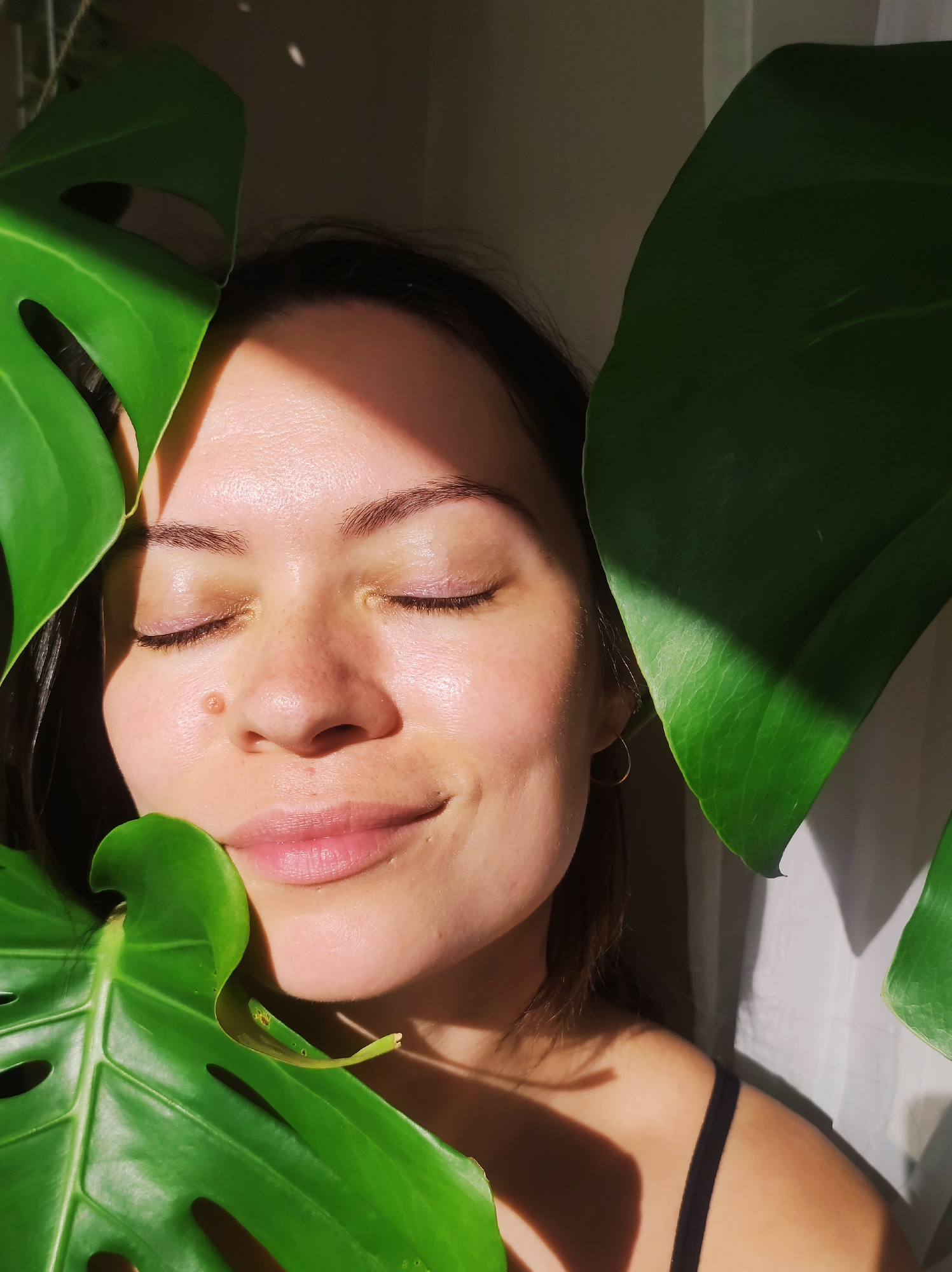 A young woman with light skin is positioned among large, green Monstera plant leaves indoors, likely in a residential space, given the domestic ambience. Her eyes are closed, and she wears a slight smile. A distinct mole is visible on the right side of her nose. She is wearing a black tank top. The background consists of a textured white wall, illuminated by sunlight. The city of Chaikovsky, Russia is not discernable from the image.