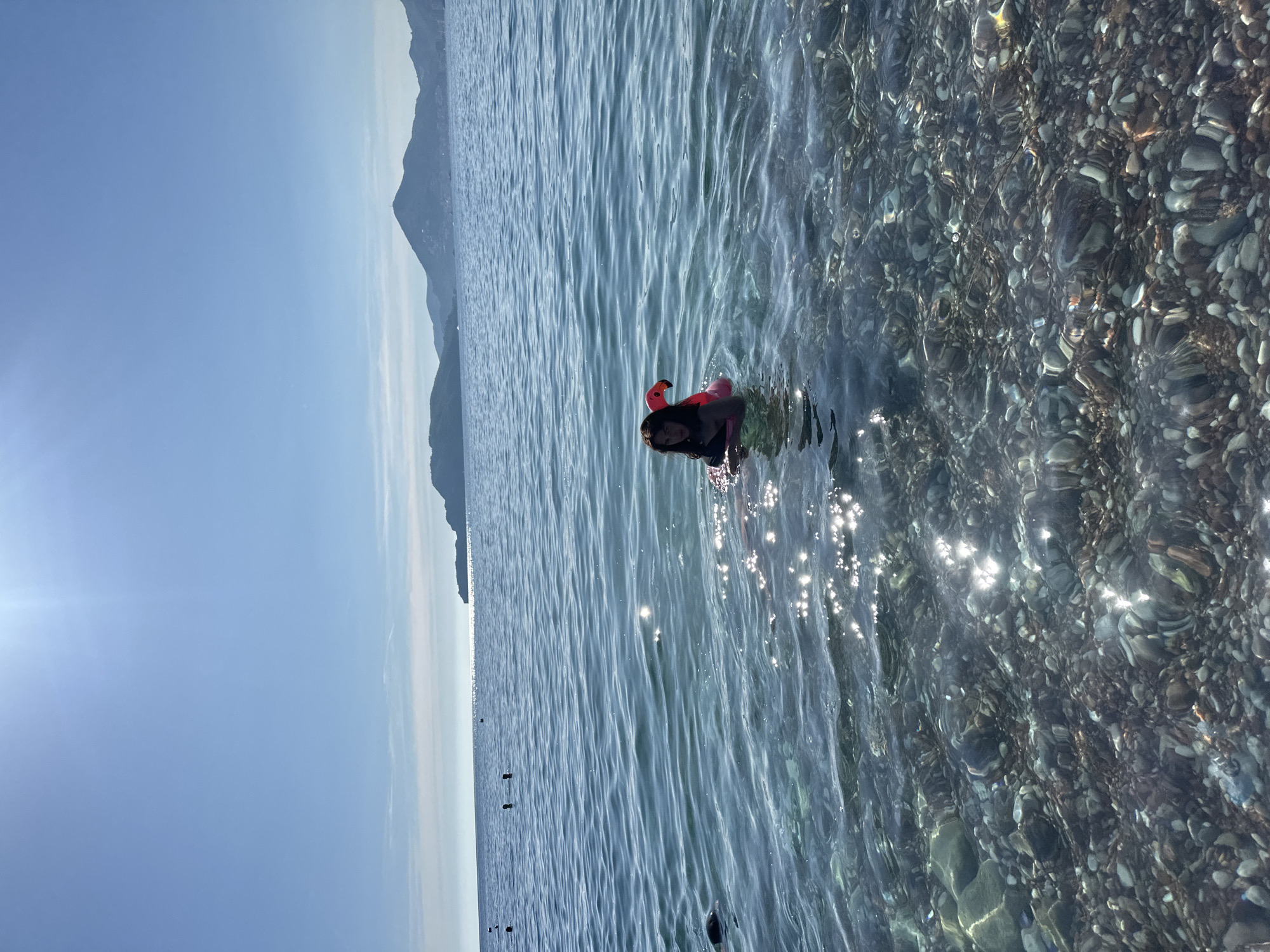 A person is snorkeling in the clear, shallow water of a pebbly beach. The person is wearing a black swimsuit and a pink snorkel mask with a red tube. Their head is mostly submerged, with only their face and the snorkel visible above the water. The sun is shining brightly, creating reflections on the water's surface. In the background, a hazy mountain range is visible under a clear blue sky, with the sun creating a lens flare on the left side of the image. The water is so clear that the pebbles on the seabed are easily seen. This scene appears to be taking place on a sunny day, likely in the late morning or early afternoon, given the angle of the sun and the brightness of the light. The overall mood is peaceful and recreational, suggesting a holiday or leisure activity at Sveti Stefan, Montenegro.