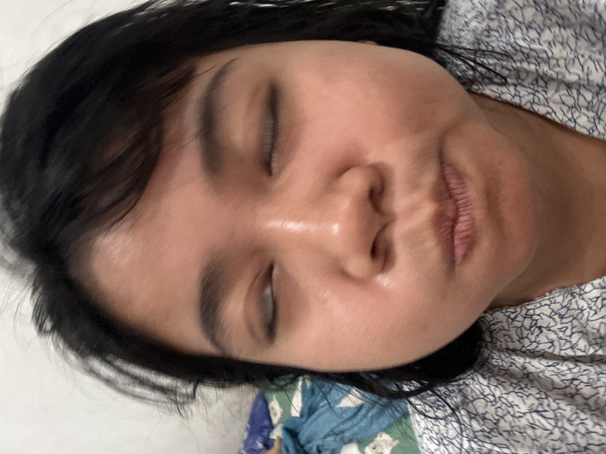This is a close-up, low-angle, indoor, eye-level, medium shot of a young woman with black hair and closed eyes. She is wearing a light-colored shirt with a dark blue, abstract pattern. Her cheeks are puffed out, and her lips are pursed. The background is blurry and shows a white wall and a blue and green patterned blanket. The lighting is soft and diffused, creating a slightly glossy sheen on her skin. The image appears to be a selfie taken in a residential setting in North Jakarta, Indonesia.