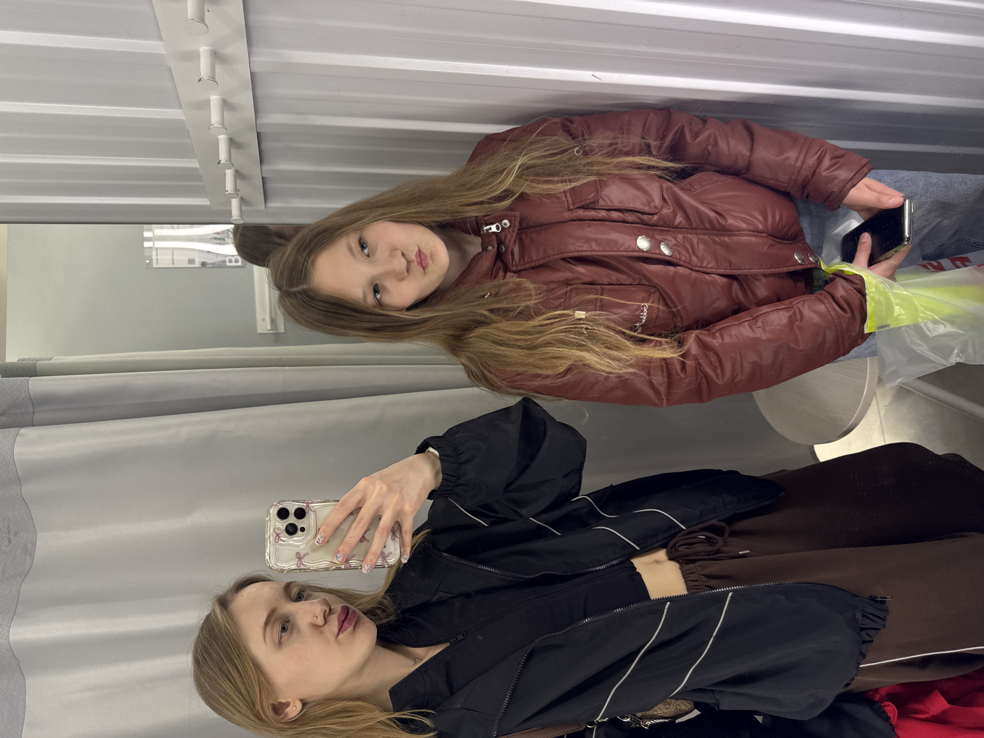 A young woman with blonde hair is taking a selfie in a fitting room using a smartphone with a decorative case. She is wearing a black jacket and brown pants. The reflection in the mirror shows another young woman with long, blonde hair wearing a maroon puffer jacket and jeans, holding a smartphone. The fitting room has gray corrugated metal walls and a white ceiling. Some clothes are visible, including a red garment. The location is presumably a retail establishment, but there is no indication that it is located in Ovoshchnoy, Russia.