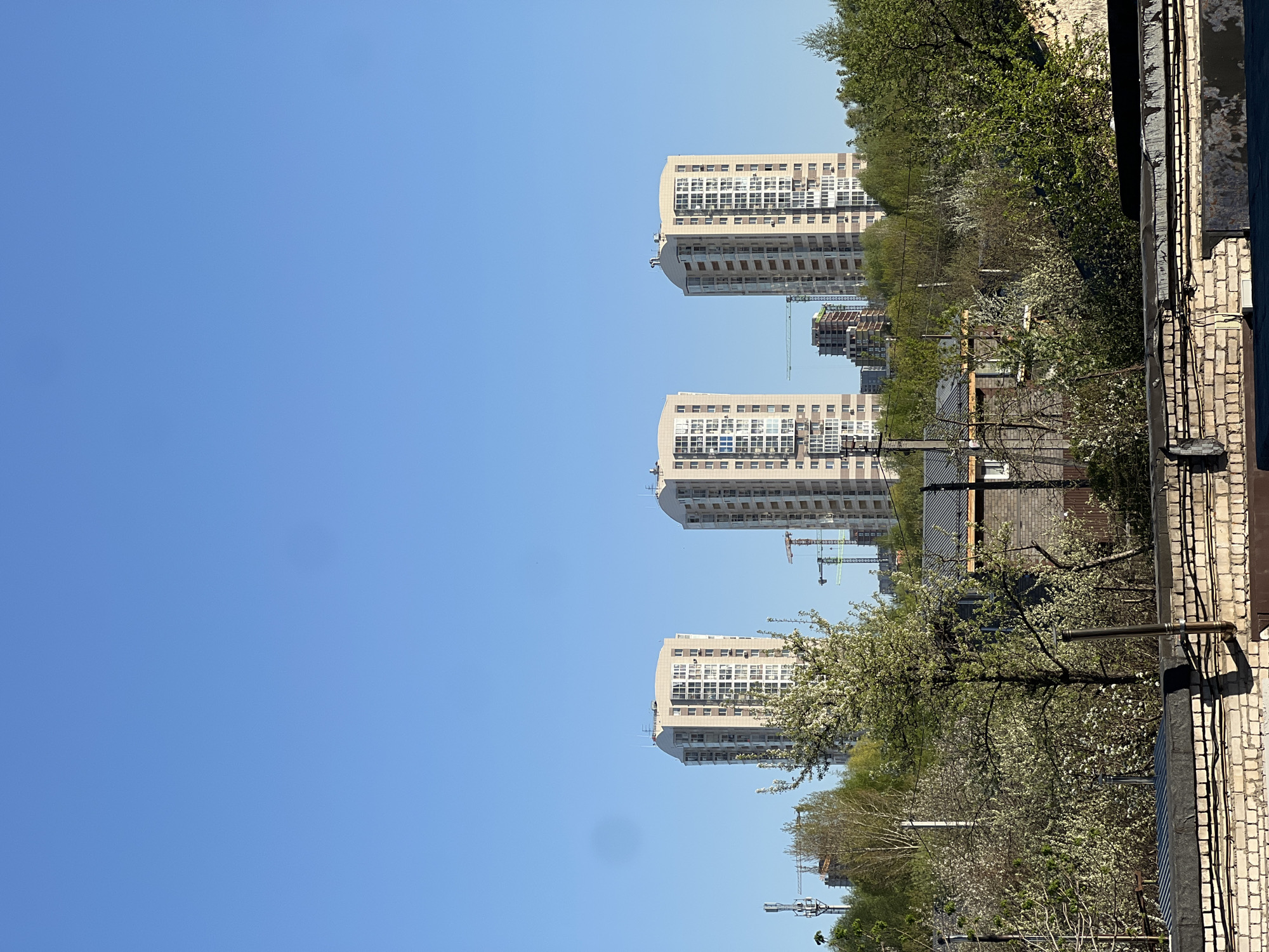 The image shows a vertical view of a cityscape in Kazan, Russia. Three tall apartment buildings dominate the skyline. Several construction cranes are visible, suggesting ongoing urban development. In the foreground, there is a tree in bloom, and a brick wall with pipes. The sky is clear and blue.