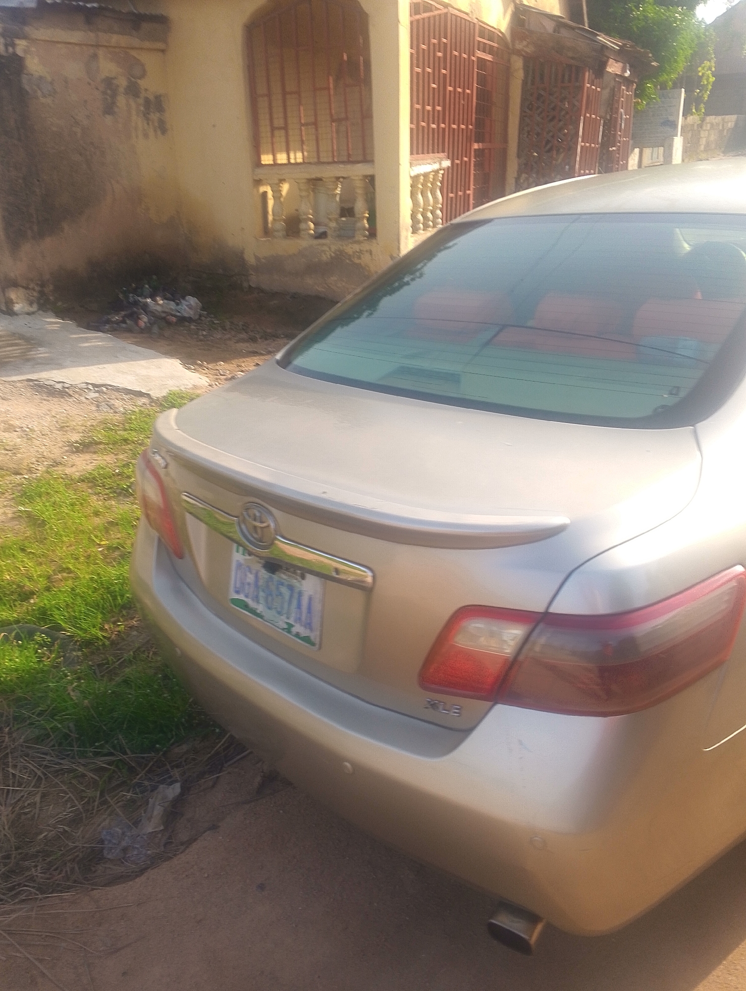 The image shows the rear passenger side of a light gold sedan parked on a dirt and grass area, likely a roadside, in Jalingo, Nigeria. The car has a spoiler on the trunk and a license plate that reads "DGA-657AA". In the background, there is a yellow building with red barred windows and a corrugated metal roof. Overgrown grass and dry vegetation are visible on the ground, along with some scattered debris. The overall impression is that of an urban or semi-urban environment.