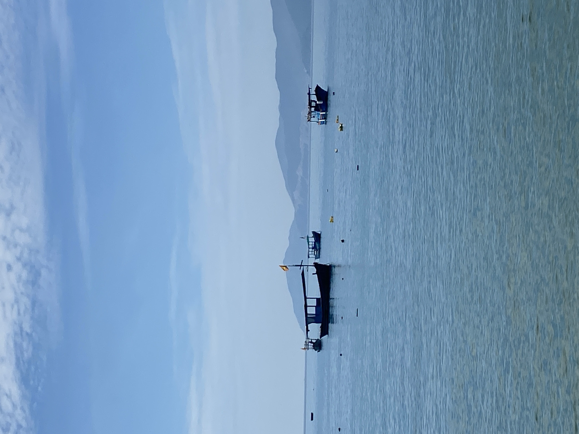 This image depicts a serene seascape with several boats anchored in calm, blue waters. In the foreground, the water is a clear, light blue with subtle ripples. The middle ground features three distinct boats. The largest boat, in the center, is dark-colored with a canopy and a yellow flag. To its left, a smaller, similar boat is also visible. Further to the right, another blue boat with a canopy is present. Scattered across the water are small yellow buoys. The background is dominated by hazy, blue mountains that stretch across the horizon. The sky above is a pale blue with scattered, wispy white clouds. The scene suggests a peaceful day at sea, likely in a coastal area. Given the presence of mountains, the clear blue water, and the style of the boats, this scene is consistent with coastal areas around Nha Trang, Vietnam, potentially a bay or a sheltered cove.