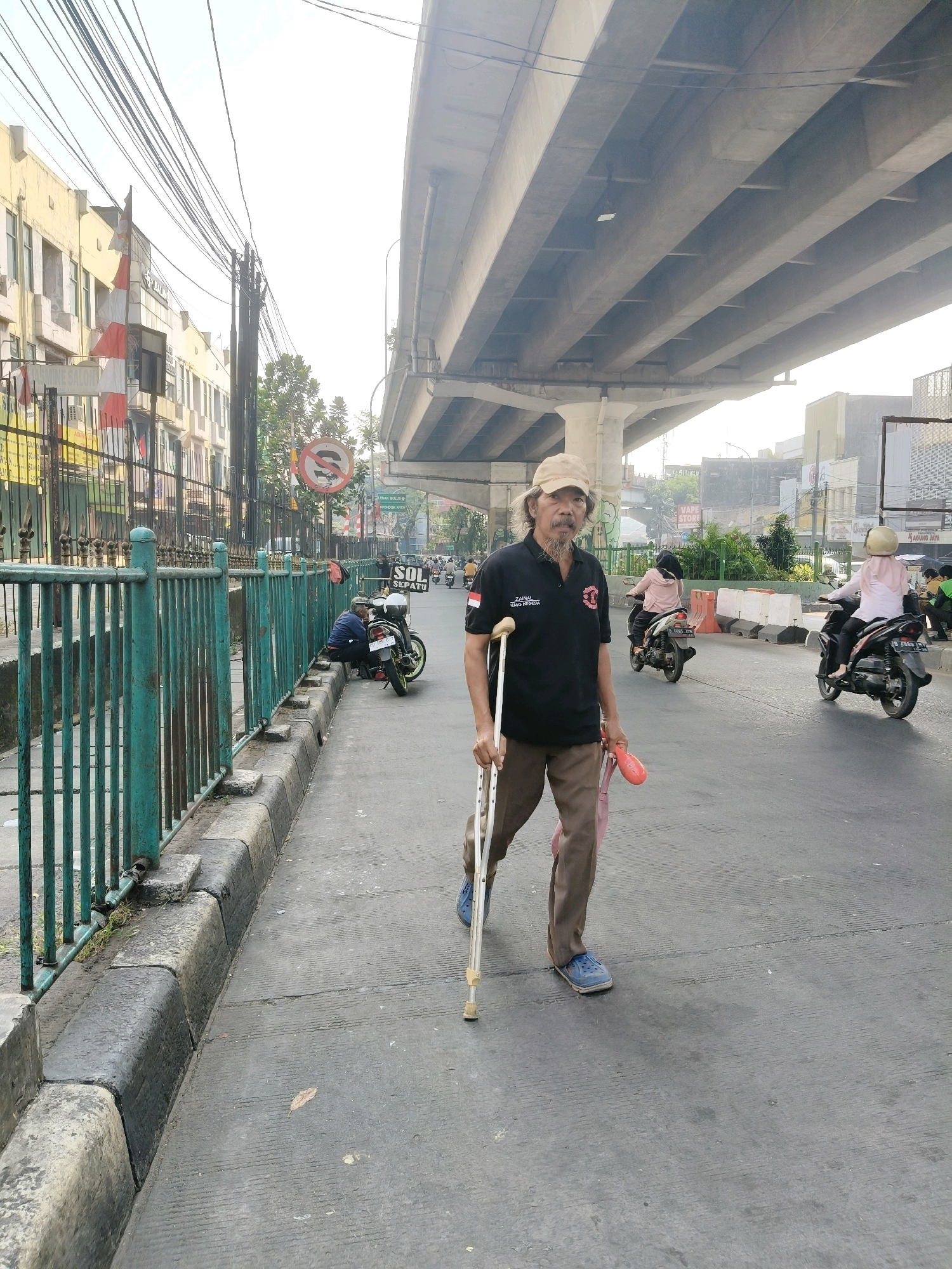 The image depicts a street scene in South Tangerang, Indonesia, under an elevated highway. A man with a mustache and a baseball cap is walking towards the camera, using a crutch in his right hand and carrying a pink object in his left. He is wearing a black polo shirt with white text on the left sleeve and a logo on the left chest, brown pants, and blue shoes. Behind him, several motorcycles are visible on the road. To the left, a green metal fence lines a sidewalk with a concrete curb. Buildings with yellow and white facades line the street behind the fence, and numerous electrical wires crisscross the sky. A "no stopping" sign with a red circle and diagonal line is posted on a pole. The overall impression is one of a busy urban environment with signs of everyday life.