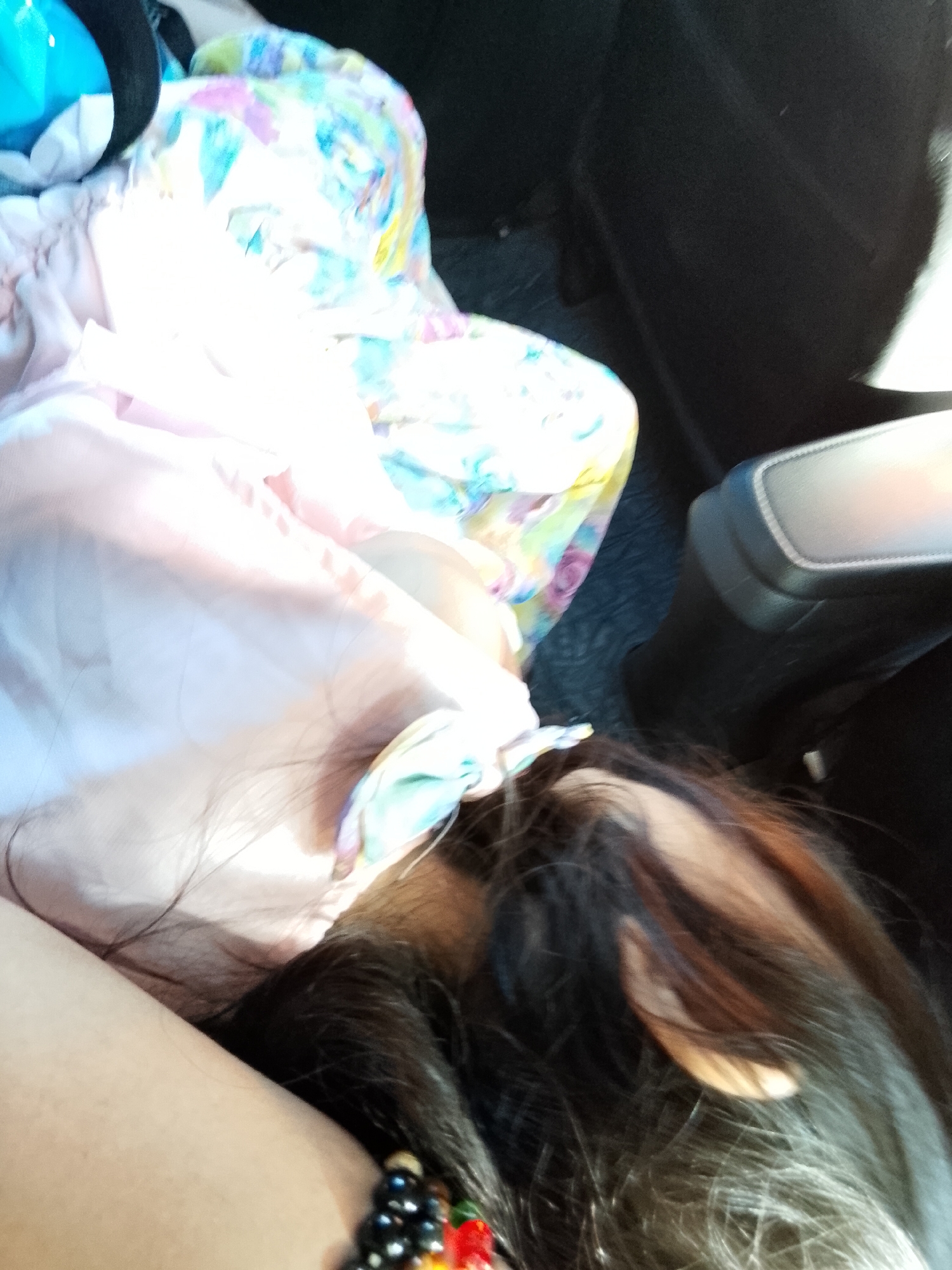 The image displays the interior of a vehicle, specifically located in Davao City, Philippines, under natural light conditions. A single individual is partially visible, primarily their head, arm, and hair. The person's dark, long hair, exhibiting some reddish-brown highlights, occupies the lower right and central portions of the frame, partially obscuring the face, with a section of the right ear visible. A fair-skinned arm is positioned in the lower-left quadrant, adorned with a beaded bracelet comprising predominantly black beads accented with smaller colorful beads (red, orange, green).
Various objects are present within the vehicle. A light pink fabric is draped across the mid-left area. Adjacent to it, towards the upper center, is a brightly colored fabric featuring a pastel floral or abstract pattern in shades of blue, pink, green, yellow, and purple. In the upper-left corner, a bright blue object with a black rim, consistent with a bottle or container, is partially visible. The vehicle's interior is characterized by dark, likely black or dark gray, upholstery and panels forming the background. A gray and black padded structure, resembling a car's armrest or center console, is positioned on the mid-right side. The overall lighting suggests direct sunlight entering the vehicle, creating bright areas and distinct shadows. The person appears to be in a resting or seated posture.