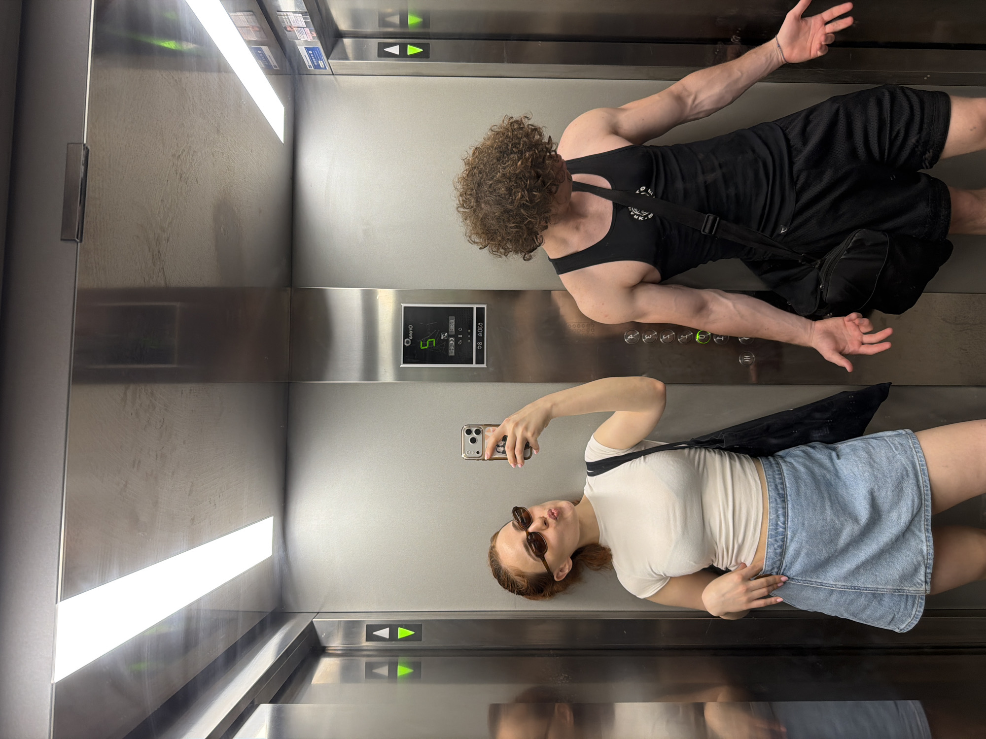A young couple is taking a selfie in an elevator. The woman is on the left, wearing a white t-shirt, denim skirt, and sunglasses. She is puckering her lips for the photo. The man is on the right, wearing a black tank top and black shorts. He has curly hair and is looking down at the floor. Both have bags slung over their shoulders. The elevator is made of stainless steel, with bright fluorescent lights on the ceiling. The elevator control panel is visible on the wall, with buttons and floor indicators. The overall impression is a casual, candid moment captured in a modern urban setting.