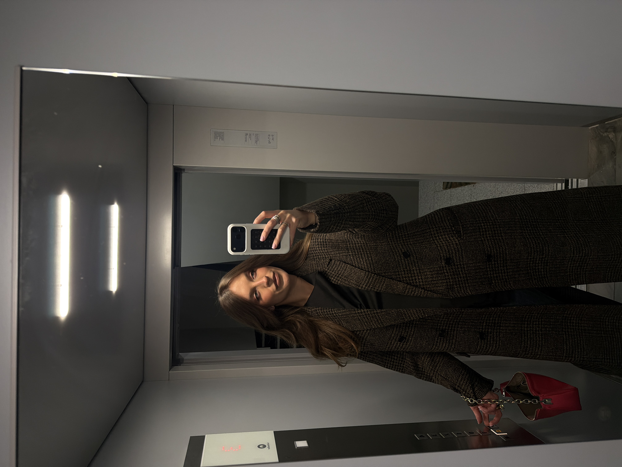 A young woman is taking a mirror selfie inside an elevator. The elevator appears to be modern, with stainless steel and white paneling. The woman is wearing a brown patterned overcoat and holding a red purse. She is pressing a button in the elevator. The location is in an urban setting. It is not possible to confirm the location is in Vozdovac Urban Municipality, Serbia from the image.