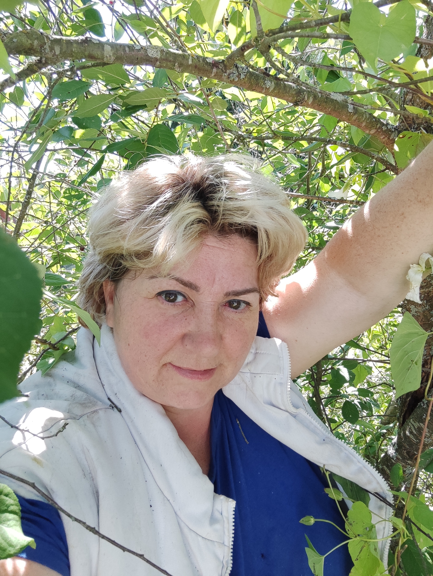 The image shows a woman in her 40s with blonde, short hair and fair skin, with freckles on her face and arms. She is wearing a blue shirt and a white zipped jacket. She is outdoors, surrounded by green foliage and tree branches. Sunlight filters through the leaves, casting shadows and creating a dappled effect. The woman is leaning against a tree, with her right arm raised and resting on a branch. Her expression is relaxed, with a slight smile. The overall scene suggests a warm, sunny day in a natural setting, possibly a garden or a park. The location is stated to be in the city of Velikii Krai, Russia.
