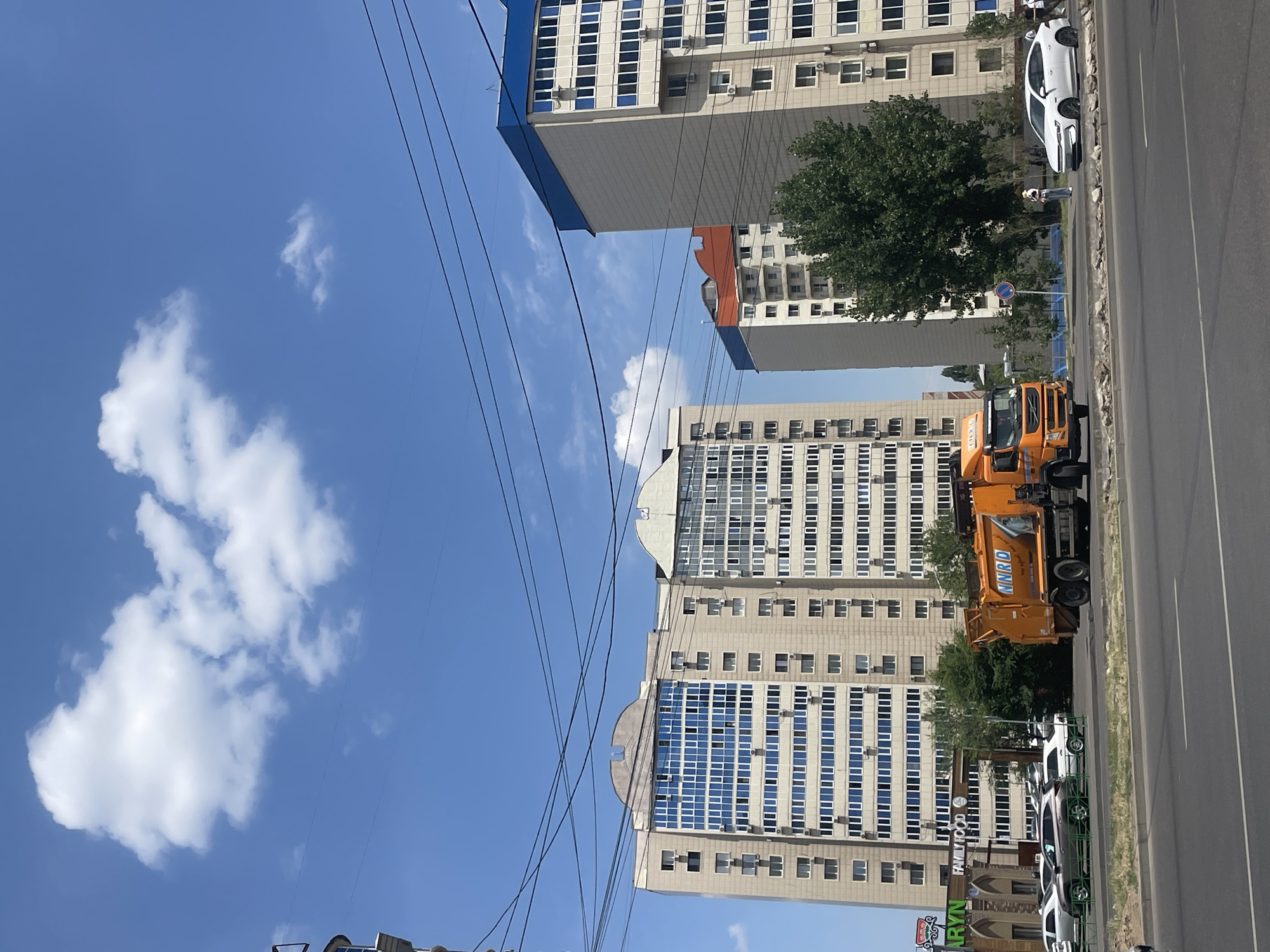 This image depicts a street scene in Almaty, Kazakhstan. The sun is shining brightly, creating a clear blue sky with scattered white clouds, suggesting it's a pleasant daytime. The composition is dominated by modern, multi-story buildings with many windows, indicating an urban environment.
In the foreground, an orange garbage truck is parked on the side of a road, its name "NRRO" visible on its side. Several cars are parked along the street, including a white sedan and a silver car. A few people are visible on the sidewalk, including one person standing near the white car, appearing to be a pedestrian. The presence of power lines strung across the sky adds to the urban feel.
Further down the street, another tall building with a blue-topped section is visible. The architecture of the buildings suggests a contemporary urban setting. The overall impression is of a typical day in a bustling city. The image is taken in portrait orientation.