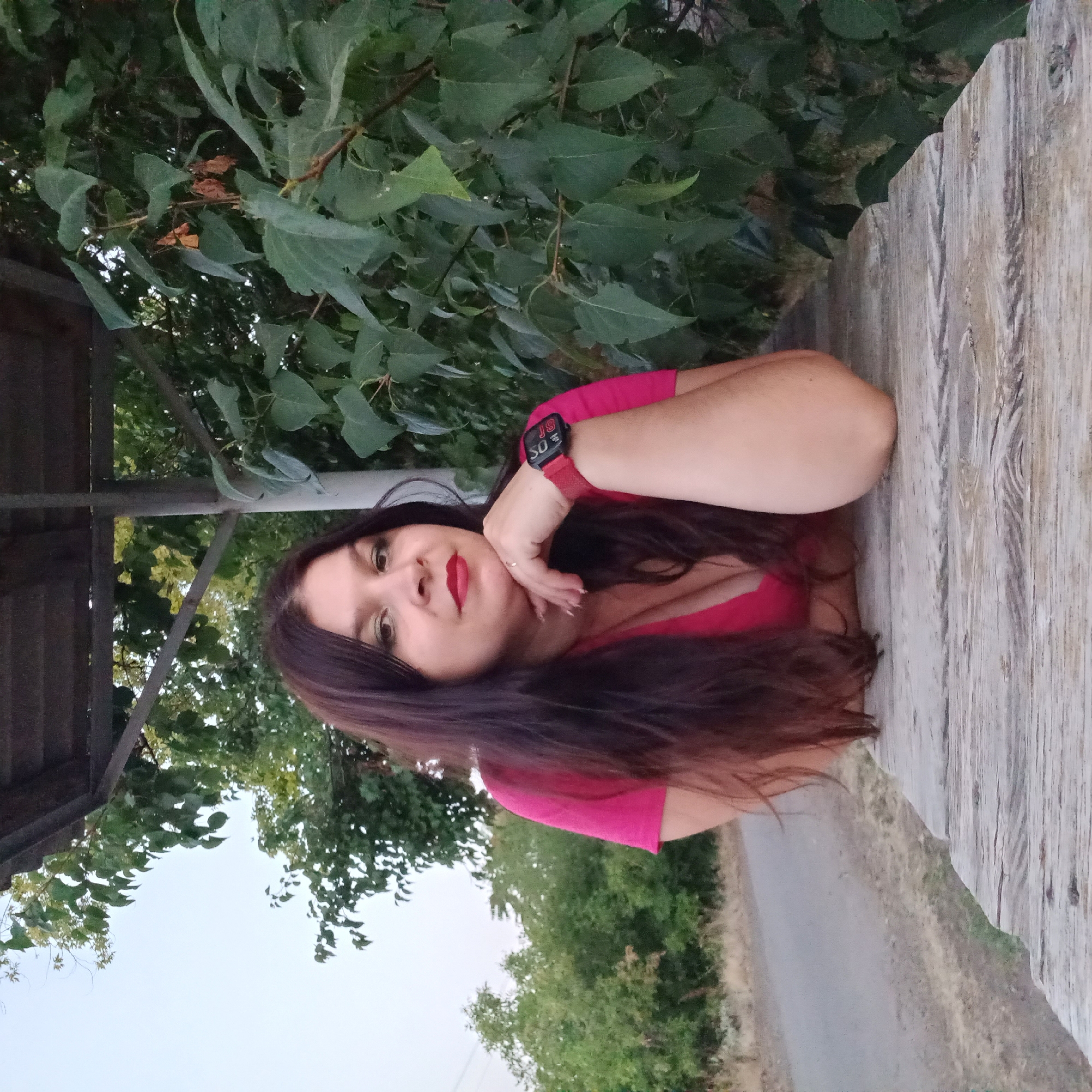 A woman with long, dark hair and green eyeshadow, wearing a pink V-neck shirt, is posing for a photo. She has red lipstick on and is resting her chin on her hand, which is adorned with a ring. A red smartwatch is visible on her wrist. She is sitting at a wooden table, with lush green foliage behind her and a road in the background. The scene is set outdoors, possibly in Berezhynka, Ukraine.