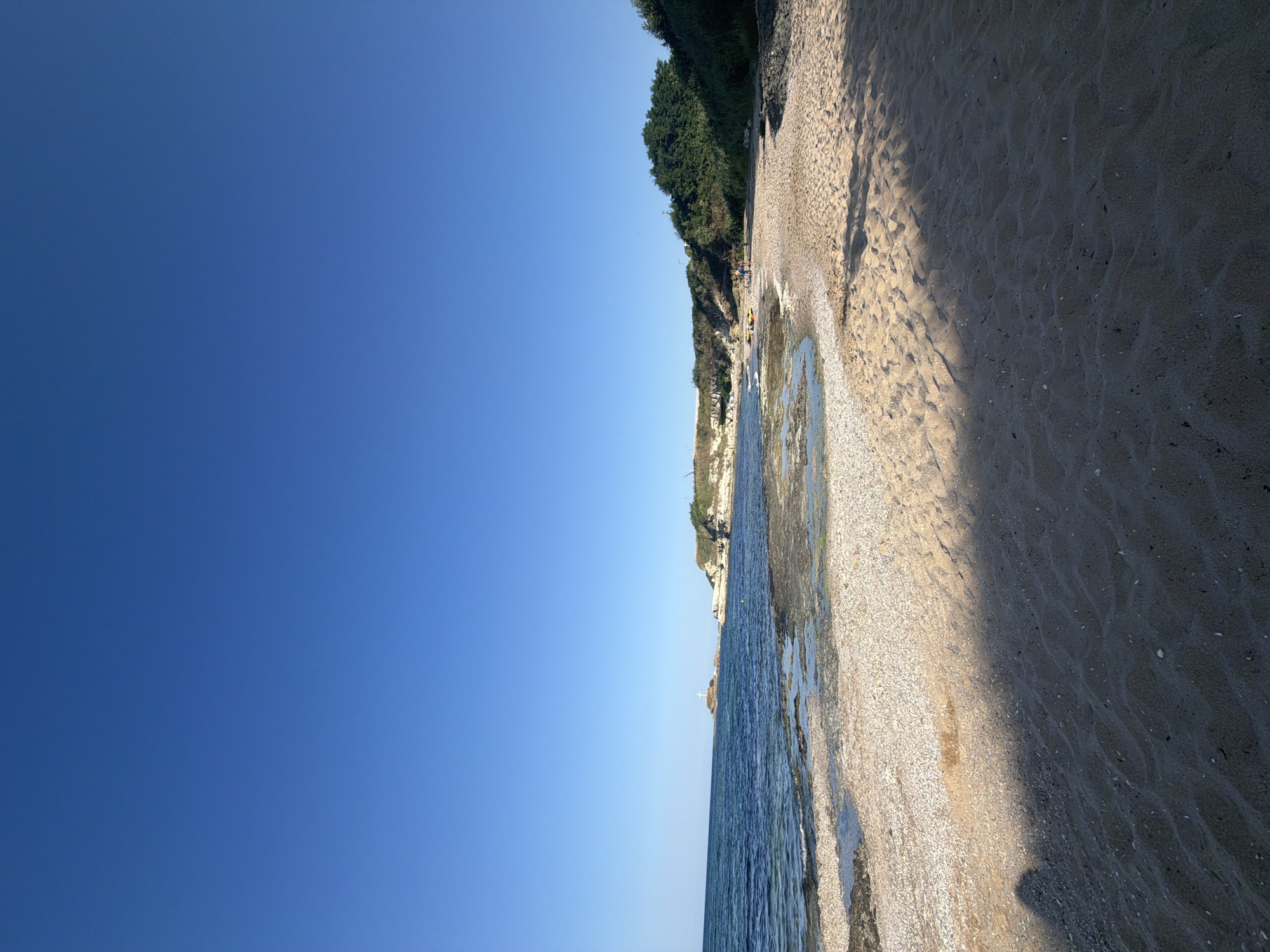 The image depicts a tranquil beach scene in Lozenets, Bulgaria, bathed in the warm glow of sunlight under a clear blue sky. The foreground is dominated by a sandy beach, with soft ripples of sand suggesting a gentle breeze. A shadow of a person is cast across the sand in the lower left corner of the frame. The midground reveals a shoreline where the sand transitions into shallow, calm water. The water is clear enough to see the rocky and shell-strewn seabed beneath. Further out, the sea stretches to the horizon, with a gentle wave pattern.
In the background, a rocky coastline curves along the sea, with patches of green vegetation and trees clinging to the higher elevations. Small white structures, possibly buildings or chapels, are visible atop the cliffs. Notably, a prominent white cross stands on a small, isolated rock formation just offshore, adding a distinctive landmark to the seascape. On the beach in the distance, a few small groups of people are scattered, some appearing to be engaged in leisure activities. Two yellow kayaks or similar watercraft are visible on the sand near some of these individuals, suggesting recreational use of the beach. The overall atmosphere is serene and picturesque, typical of a summer day in a coastal Bulgarian town.