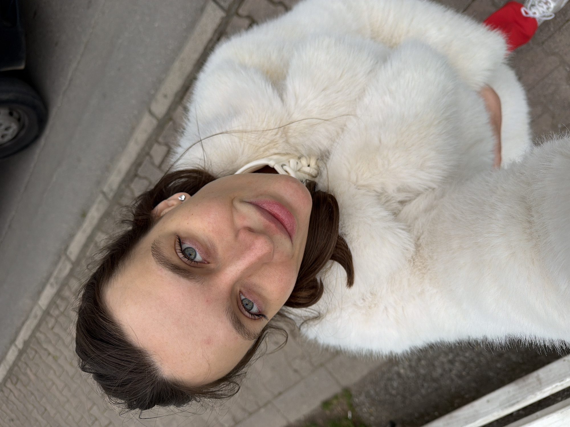 A woman is seen from an overhead perspective, wearing a large, white fur coat. Her dark hair is pulled back, and she has light eyes. A small earring is visible in her left ear. The background includes a sidewalk, a curb, and a portion of a vehicle with a visible tire. A red object is visible in the upper right corner. The location is presumed to be Budva, Montenegro.
