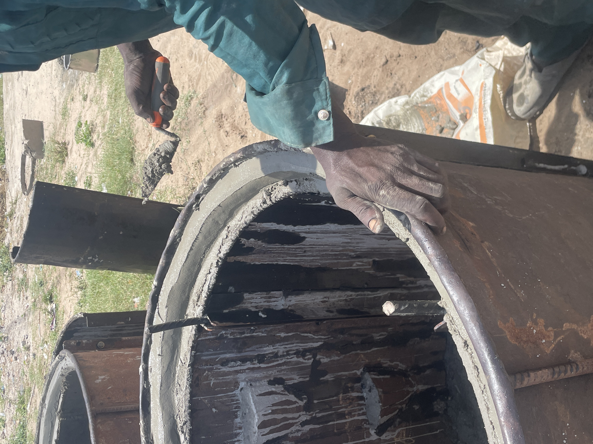 The image provides a high-angle, close-up view of a person engaged in construction work outdoors in Badagry, Nigeria. The scene is brightly lit by daylight, indicating clear weather.
The primary subjects are the hands and forearms of a worker, clad in a teal or dark green long-sleeved work shirt with a cuff visible on the right arm. Their dark-toned hands are actively involved in working with cement.
In the left foreground, one hand holds a small hand trowel with an orange and black handle, laden with wet cement or mortar. This hand appears to be applying the material. Simultaneously, the other hand, positioned towards the right, is seen smoothing fresh cement along the rim of a large, circular metal form. This form is likely a mold for a concrete pipe or culvert, with steel reinforcing bars (rebar) visible within its interior. Another similar circular form is partially visible behind the one being worked on.
The ground beneath and around the activity is sandy, light-brown soil with scattered patches of sparse green grass and some dry vegetation. In the background, a white and orange bag, potentially a cement bag given the context, lies on the ground next to a partially visible light-colored shoe or boot. Faint orange lettering on the bag is visible, although not fully legible.
The activity clearly depicts the manual process of casting or finishing a concrete element, showcasing the application and shaping of cement. The overall setting is an unpaved outdoor area typical of a construction site or a pre-casting workshop.