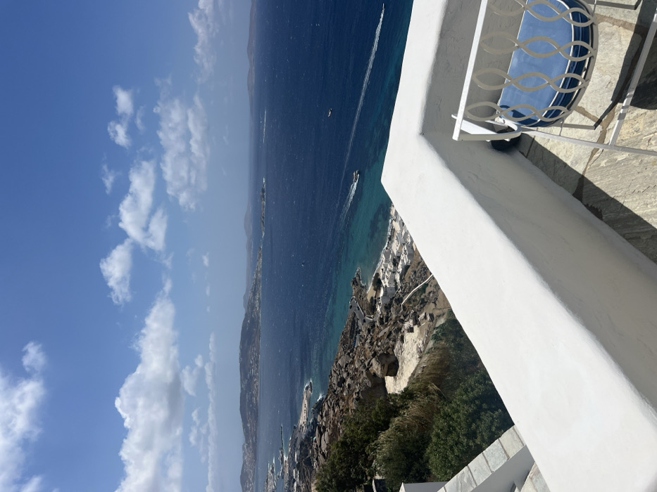 A panoramic daytime view from an elevated balcony in Tourlos, Mykonos, Greece, overlooks the Aegean Sea and the adjacent coastline under a clear blue sky with scattered cumulus clouds. In the immediate foreground, a white, L-shaped balcony railing and light-colored stone flooring are visible, with a white metal chair featuring a blue seat cushion positioned in the lower right corner. Below the balcony, a steep, rocky hillside covered with sparse green vegetation descends towards the shoreline. Small, white-washed buildings are clustered along the rugged coast, where the water is a clear turquoise. Further out, the sea is a deep blue, with at least two small watercraft observed moving, leaving distinct white wakes. Distant landmasses are visible on the horizon. No individuals are present in the scene.