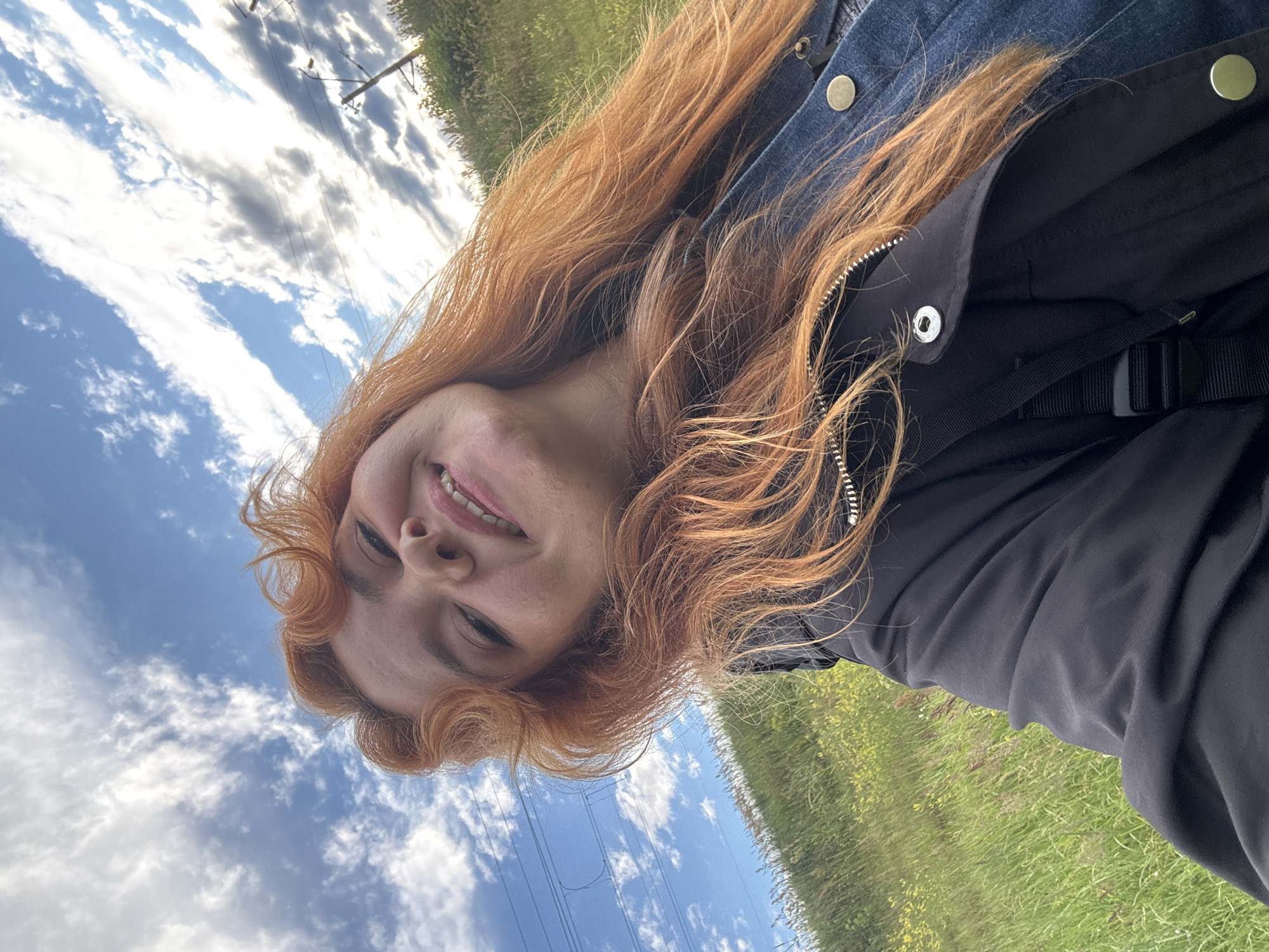 A young woman with long, wavy, reddish-brown hair smiles at the camera in a selfie. She is wearing a denim jacket over a black garment, possibly a t-shirt or a jacket with a zipper. Her hair is loose and partially obscures her face. The background is a bright, cloudy sky, with some green grassy fields visible to the right. There are also hints of power lines and poles in the distance. The lighting suggests it is daytime, and the weather appears to be partly cloudy. The woman's expression is joyful and relaxed. The image has a slightly tilted perspective, as if taken from a low angle or by someone holding the camera at an unusual position. The visible part of her face includes her smiling mouth and part of her nose and eyes. There is no visible text in the image.