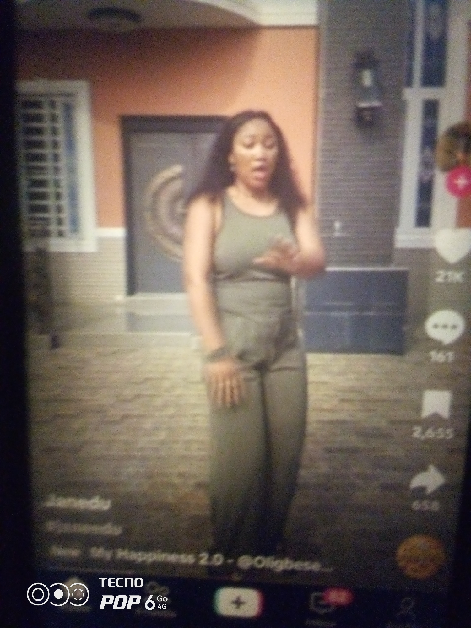 The image is a screenshot of a mobile phone screen, likely from a social media application like TikTok. It features a woman standing in front of a building. She is wearing a sleeveless olive-green top and matching pants. Her hair is dark and extends past her shoulders. She is looking towards the camera with her mouth open, suggesting she is singing or speaking. In the background, the building appears to be a home with a large, dark-colored door with decorative circular elements. Windows with white grilles are visible on the left side of the building. The ground in front of the building is paved with textured material. On the right side of the screen, there are icons indicating likes, comments, shares, and possibly other social media interactions, with numbers such as "21K" and "161". At the bottom of the screen, there is a logo for "TECNO POP 6 4G". Text overlays on the bottom left of the image include "Janedu", "#janedu", and "New My Happiness 2.0 - @Oligbese...". The image's resolution is low, with a grainy texture, suggesting it was taken under poor lighting conditions or with a low-quality camera. There is no visual information to determine the specific location within the city of Howayi, Nigeria.